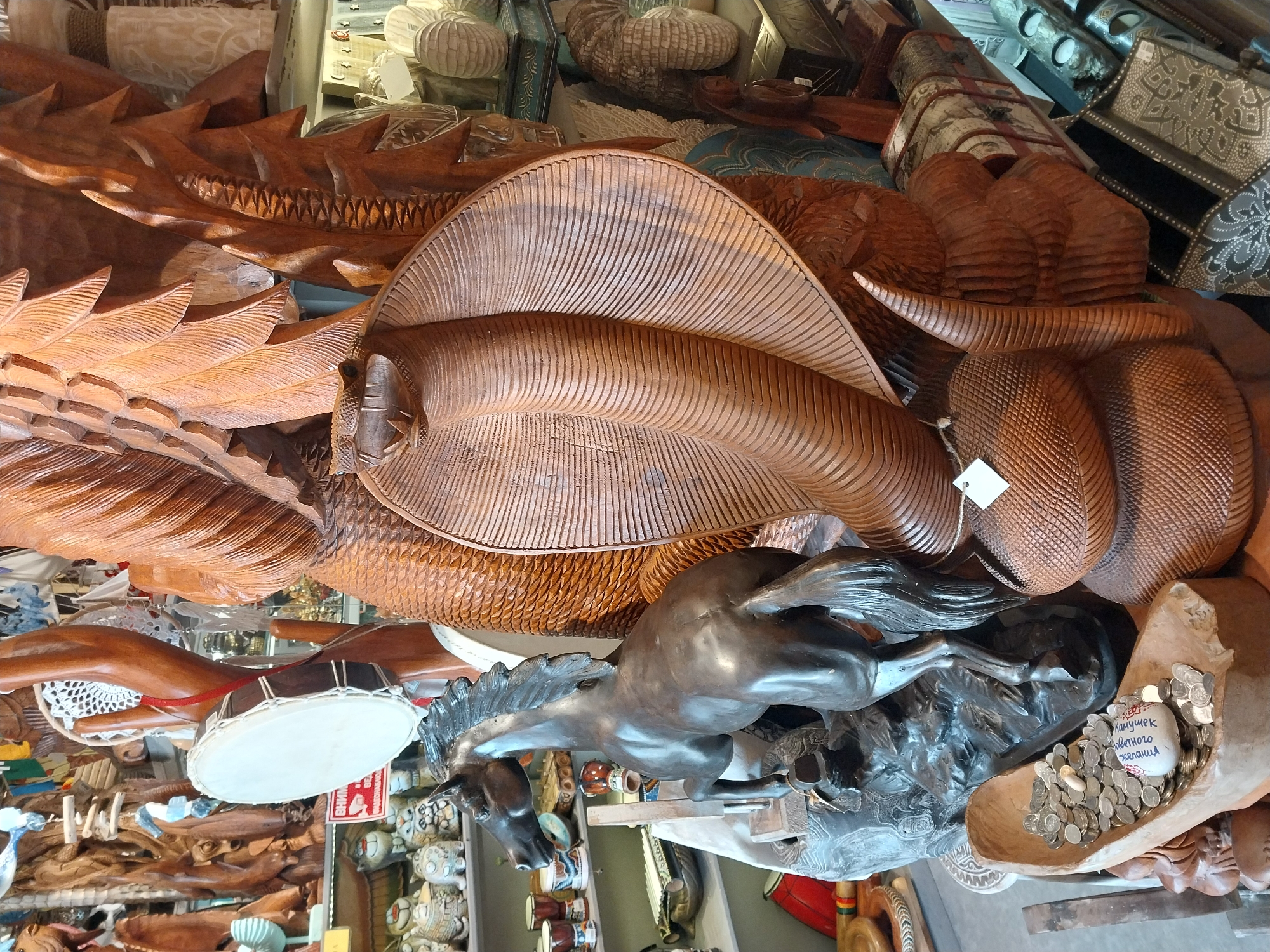 This media file displays a richly detailed collection of artisanal wooden carvings and other handcrafted objects, likely from a souvenir shop or market in Sochi, Russia. The scene is dominated by a large, intricately carved wooden piece resembling a stylized marine creature, possibly a fish or ray, with detailed scales and fin-like structures. Adjacent to it is another substantial wooden carving, appearing as a coiled serpentine form, possibly a snake, characterized by a textured, scaly surface.
Towards the foreground and slightly below the central wooden carvings, a dark-colored statue of a horse in a dynamic, galloping pose captures attention. Below the horse, a wooden trough-like container holds numerous coins, and within these coins rests a small, white, irregularly shaped object. This object bears Cyrillic text: "ПАНУЧЕК ОВЕНЧАНОГО ЖЕЛАНИЯ," which roughly translates to "Little piece for a crowned/fulfilled wish."
Other items visible include a small drum with a white head and dark rim, suspended nearby. The background is filled with various other handcrafted items, including smaller carved wooden pieces, what appear to be ceramic figures, and decorative boxes. The predominant color palette is natural wood tones, showcasing the varied grains and finishes of the materials. The overall impression is one of skilled craftsmanship and a diverse array of unique, culturally inspired souvenirs. No people or specific activities beyond the display of goods are visible.
