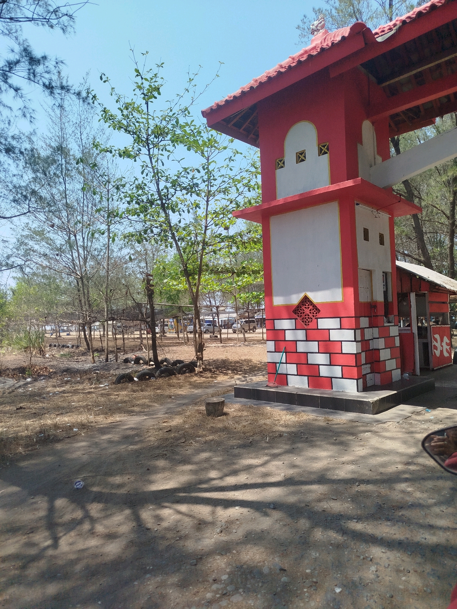 This is a full, outdoor, eye-level, vertical, daylight shot of a red and white structure that appears to be an entrance or gate at a beachside location in Setrojenar, Indonesia. The structure has a red tiled roof, a red base, and white walls with decorative red accents. In front of the structure, a green broom leans against the wall. To the right of the main structure, a smaller, red-and-white, shed-like building is visible. The surrounding environment is dry and sandy, with scattered trees, some with sparse green leaves and others bare. In the background, a beach area is visible with parked vehicles, suggesting a public gathering spot. The ground in the foreground is dirt and sand, with shadows from trees cast across it. There are some old tires and debris scattered on the ground. A person's leg, wearing a red garment, is visible in the bottom right corner, suggesting the photo was taken from a seated position.