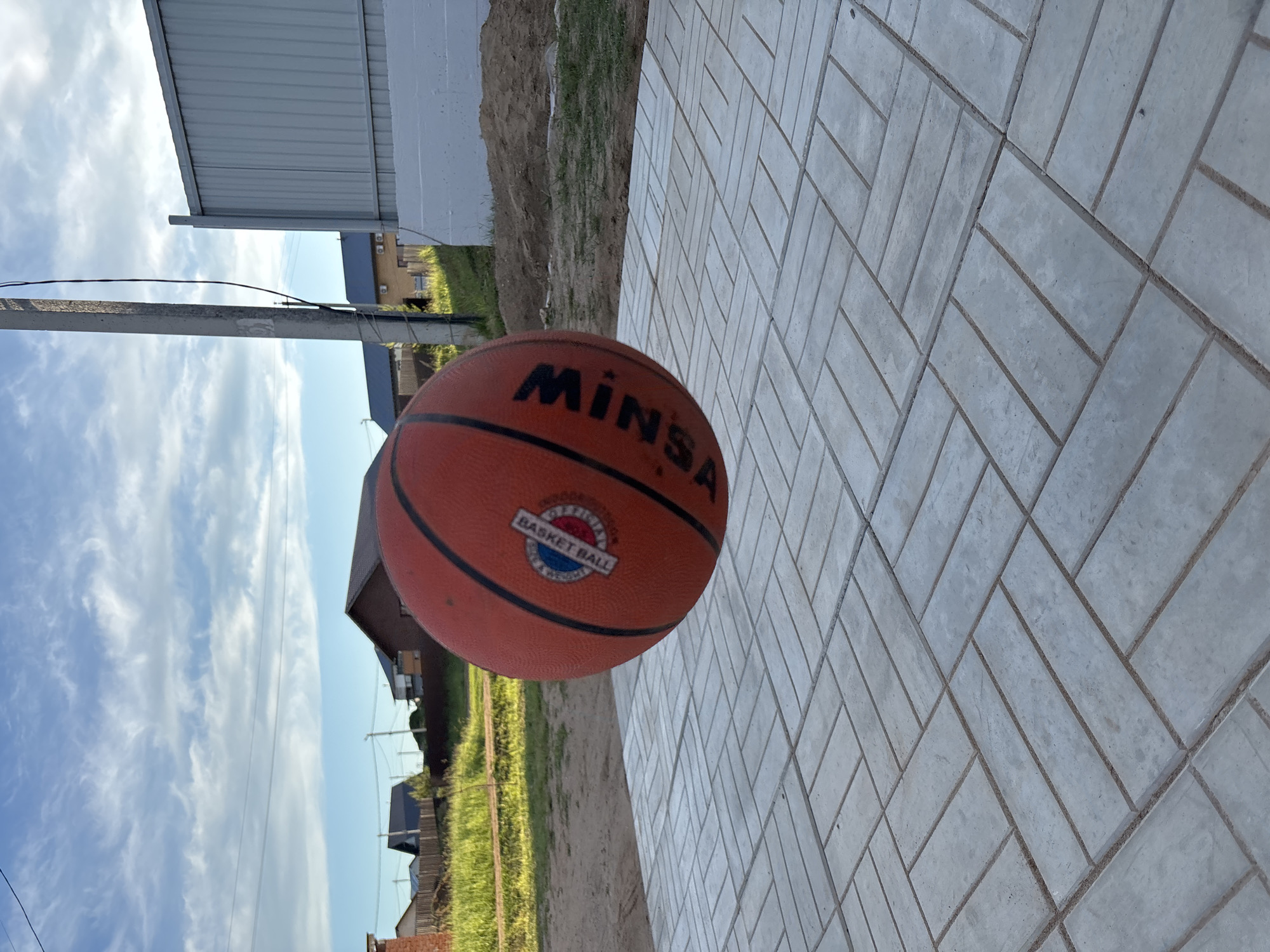 An orange basketball, branded "MINSA" and featuring a circular "BASKET BALL B.C." logo, rests prominently in the foreground of the image. The ball is positioned on a light-colored paved surface composed of rectangular blocks arranged in a distinct herringbone pattern. Adjacent to the paving, there is an area of loose dirt and green grass.
In the background, a residential or industrial building with a silver corrugated metal roof and a white facade is visible, along with other structures featuring dark roofs. A utility pole with power lines extends vertically across the midground. The scene is set outdoors under a bright blue sky adorned with scattered white, wispy clouds, indicating daytime. The overall environment suggests a suburban or semi-rural setting in Chaikovsky, Russia. No people are present, and no specific activity is taking place.