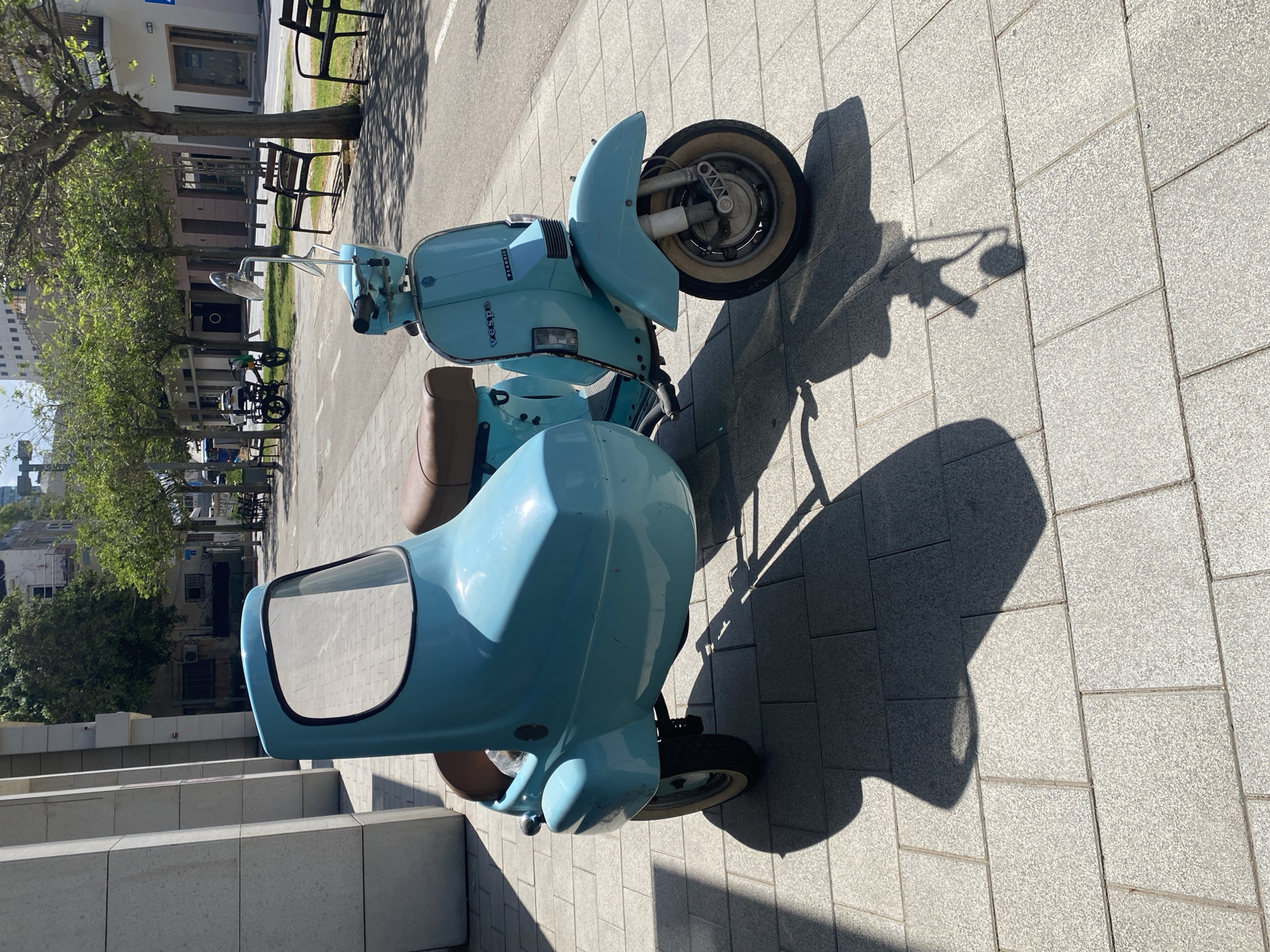 The image, rotated 90 degrees clockwise, prominently features a light blue vintage Vespa scooter with a matching sidecar, parked on a paved outdoor area under bright sunlight. The scooter, facing what would be the top of a correctly oriented frame, has a brown seat and chrome accents, with the words "Vespa" and "Piaggio" visible on its side panel. Its front wheel has a distinct white wall tire. The sidecar, positioned to the left in the rotated view, is also light blue and includes a rectangular window opening.
Long, sharp shadows from the scooter, sidecar, and surrounding elements are cast across the light grey rectangular pavement tiles, indicating a clear, sunny day. To the right of the scooter, a narrow strip of grass with green-leaved trees runs parallel to the pavement. In the background, an urban environment unfolds with various buildings, some appearing older and others more contemporary with glass fronts. Benches and a parked bicycle are also visible further back, suggesting a public space or thoroughfare. The scene depicts a stationary vehicle in a vibrant city setting, consistent with the provided location context of Tel-Aviv, Israel.