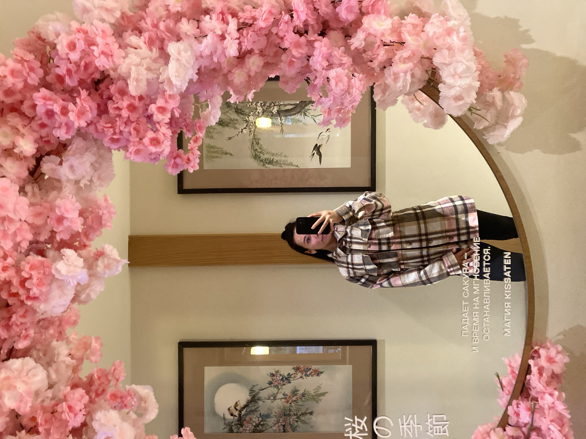 A woman takes a picture of herself in a mirror decorated with pink flowers. The mirror is round and hung on a wall between two framed pieces of artwork depicting birds and branches. Above the top artwork, there is a wooden panel, and some text is visible in the mirror. The text translates to "Sakura falls - and time for a moment stops. The magic is Kissaten." The location is Krasnodar, Russia.