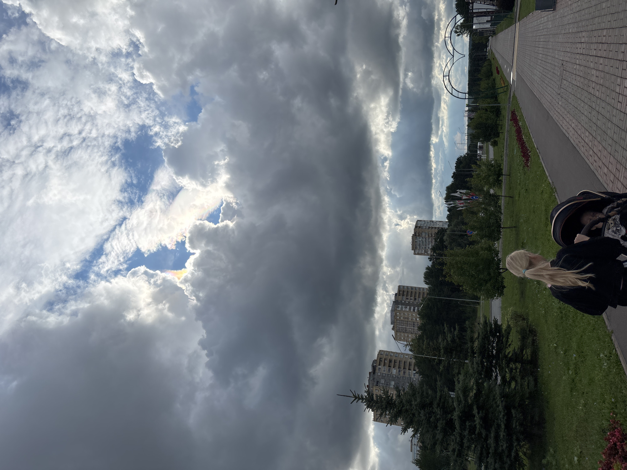 In Naberezhnye Chelny, Russia, on a cloudy day with breaks in the clouds revealing a glimpse of blue sky and a faint rainbow, a person with blonde hair in a ponytail pushes a stroller down a paved walkway. In the background, several multi-story apartment buildings stand amongst lush green trees. A large evergreen tree is positioned to the left of the frame, in front of one of the buildings. The walkway is lined with flowerbeds and leads towards a large decorative arch shaped like a heart. The scene appears to be a park or public space within the city.