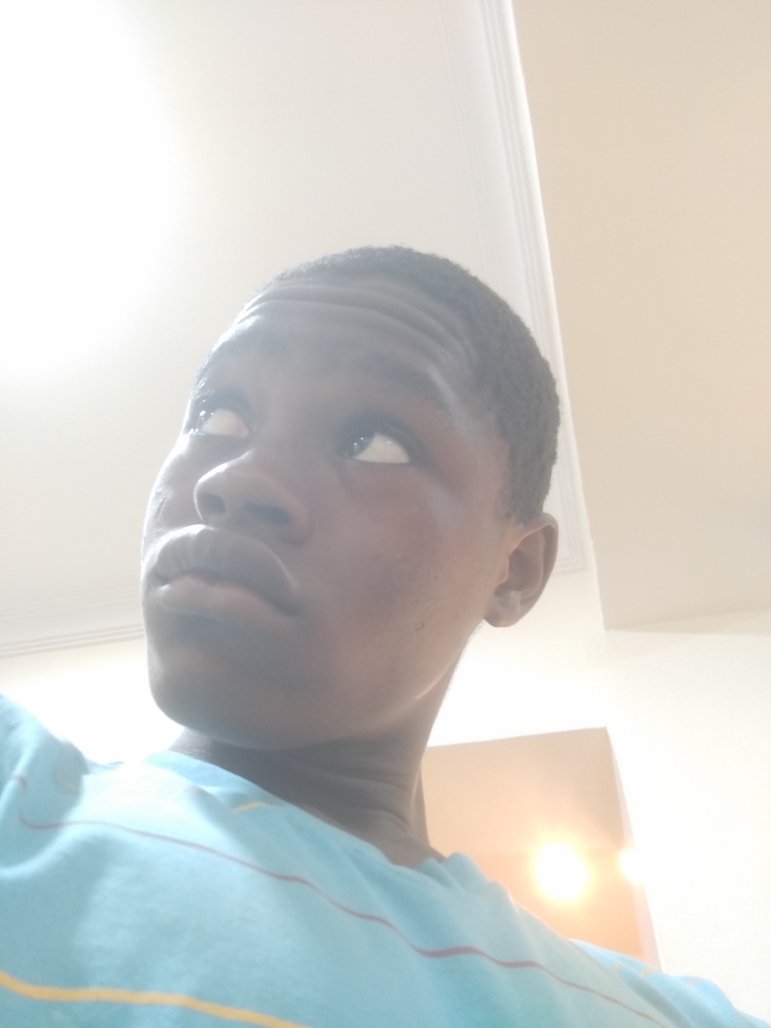 A low-angle, close-up photograph shows a Black male with short, dark hair and brown skin. He is looking up and slightly to the left. He has full lips, a broad nose, and visible eyelashes. He is wearing a light blue t-shirt with thin, horizontal stripes of yellow and red. The background is mostly blurred and overexposed, showing a light-colored wall and a glimpse of a bright light source, possibly a ceiling light, but no identifiable architectural features linking to Shangisha, Nigeria. The photo is taken indoors.