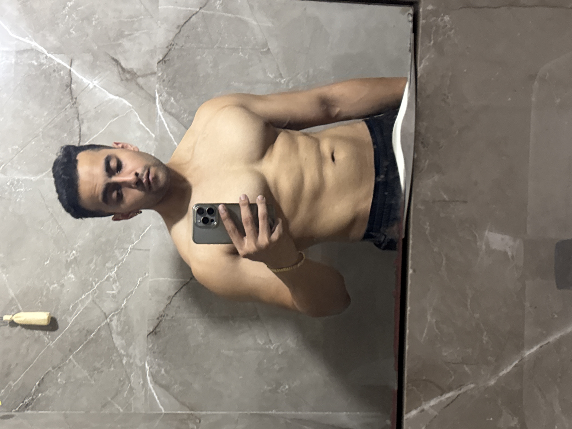 A shirtless, muscular man takes a mirror selfie in what appears to be a bathroom or dressing area. He is reflected in a large mirror, holding a dark-colored smartphone (likely an iPhone) with his right hand. His body is positioned to display his well-defined abs and chest muscles, with a slight tilt to his head and a serious expression. He wears dark underwear or shorts, and a thin golden-colored bracelet is visible on his left wrist.
The background seen in the mirror's reflection is a wall adorned with grey marble or marble-patterned tiles, characterized by distinct white vein patterns. A white sink or counter edge is visible on the right side of the reflection. In the lower-left corner, a small, yellow bottle, possibly shampoo or lotion, is reflected on a surface. The lighting is soft and even, accentuating the man's physique. There is no visible text in the image. The location is contextualized as Kolkata, India.