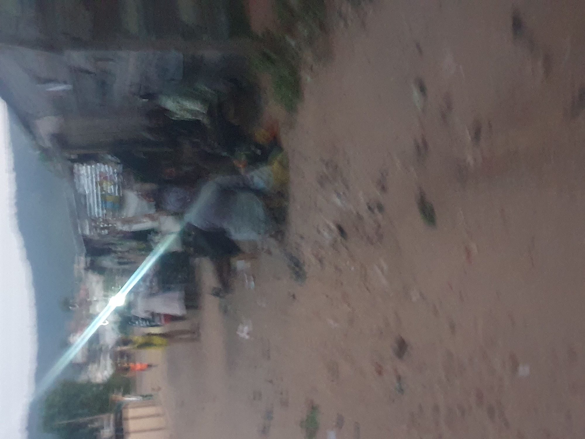 The image depicts an outdoor scene in Lokoja, Nigeria. A dirt road runs through the frame, with structures and vegetation visible on either side. People are present, some walking along the roadside. There is no clear definition of specific objects, actions, or interactions, but the general impression is of a local street scene in the city.