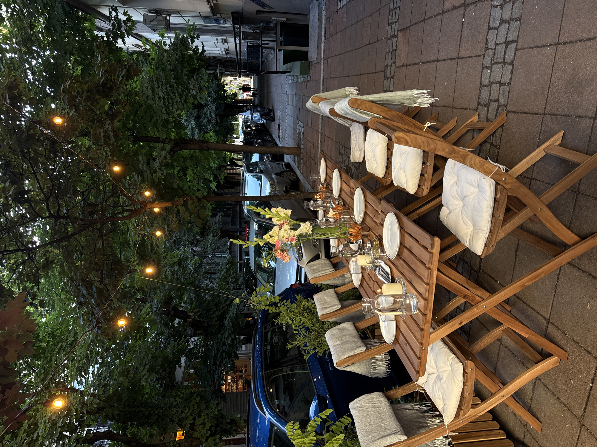 The image depicts an outdoor dining setup on a sidewalk in Belgrade, Serbia. A long, wooden table is set for a meal with white plates, silverware, glasses, and candles. A vase of colorful flowers sits in the center of the table. Several wooden chairs, each with a white cushion and a light grey throw blanket draped over the back, surround the table. String lights are strung overhead, casting a warm glow.
In the background, a blue car is parked on the street next to the sidewalk. Other cars and a scooter are also visible, along with a pedestrian walking with two dogs further down the street. Buildings line the opposite side of the street. The environment appears to be a street with mature trees providing shade.