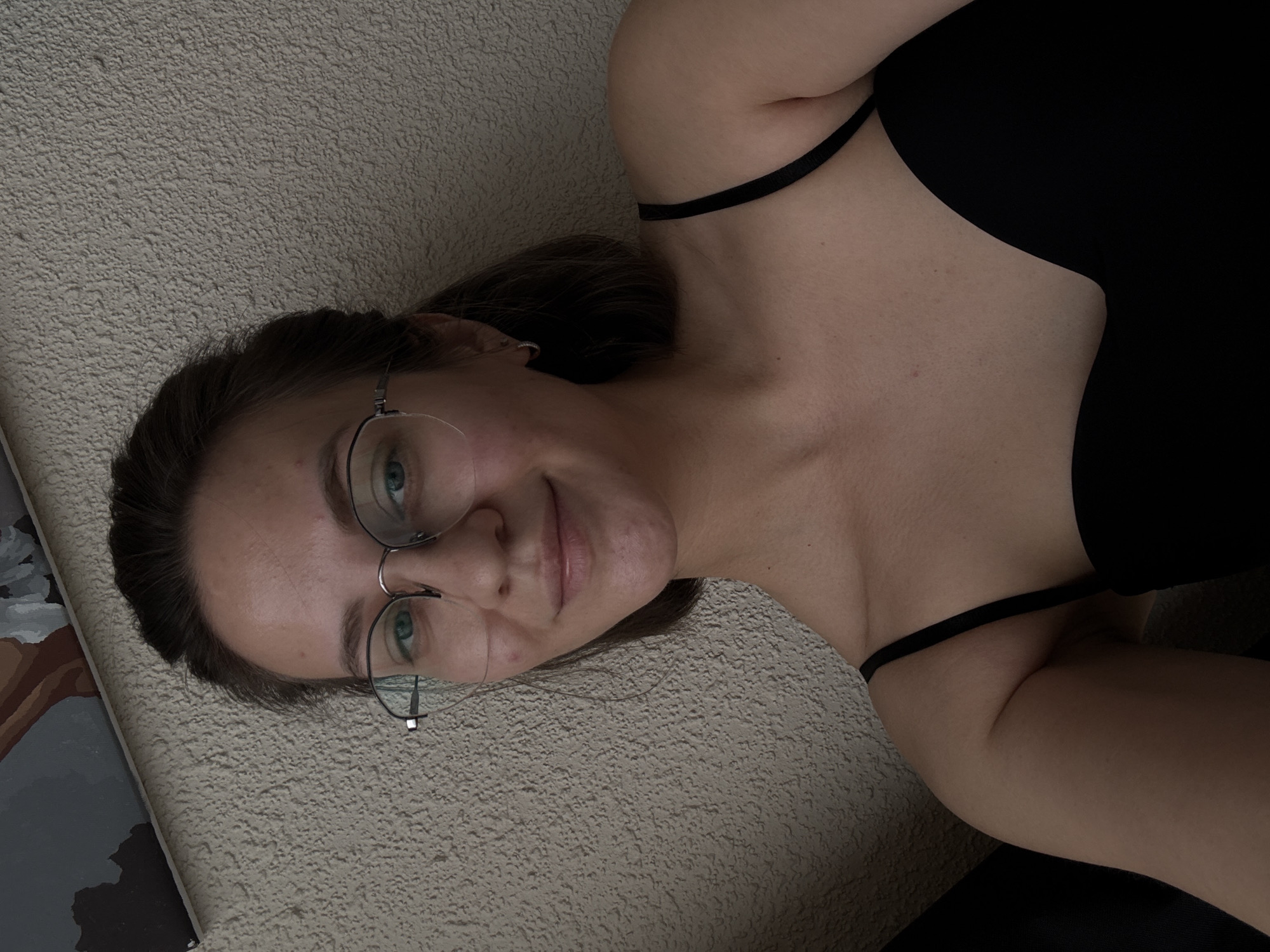 A young woman with light brown hair, wearing clear-rimmed glasses and a black camisole, smiles softly as she looks directly at the camera. Her hair is pulled back loosely, with strands framing her face. She is positioned in what appears to be an indoor setting, leaning against a textured, light-colored wall. To the upper left of the image, a portion of a painting or framed artwork is visible. The scene is lit from the front, casting subtle shadows on her face and body. There are no other individuals or significant objects in the frame.