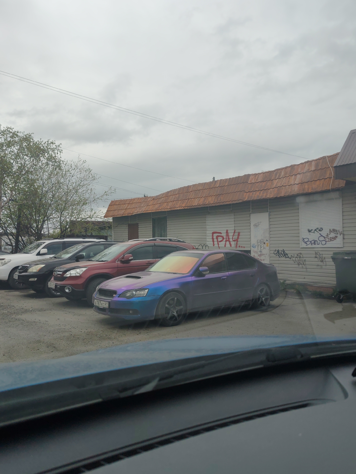 An exterior, eye-level, medium shot captures a parking lot scene under a cloudy sky. The foreground is dominated by the dark blue dashboard of a vehicle, with a portion of the windshield visible at the top. In the midground, several cars are parked in a line. The closest car is a purple Subaru Legacy, its front end angled towards the right. Behind it is a red Honda CR-V, followed by a black SUV, and then a white SUV. The cars are parked on a gravel surface. To the right of the cars, a light-colored, single-story building with a brown, tiled roof is visible. Graffiti covers the building's facade, including red lettering that reads "ГРАЧ..." on the left side. A large, dark green trash bin is positioned to the right of the building. In the background, a few trees with sparse green foliage stand to the left of the building, and power lines crisscross the sky. The overall scene depicts a somewhat weathered and urban environment.