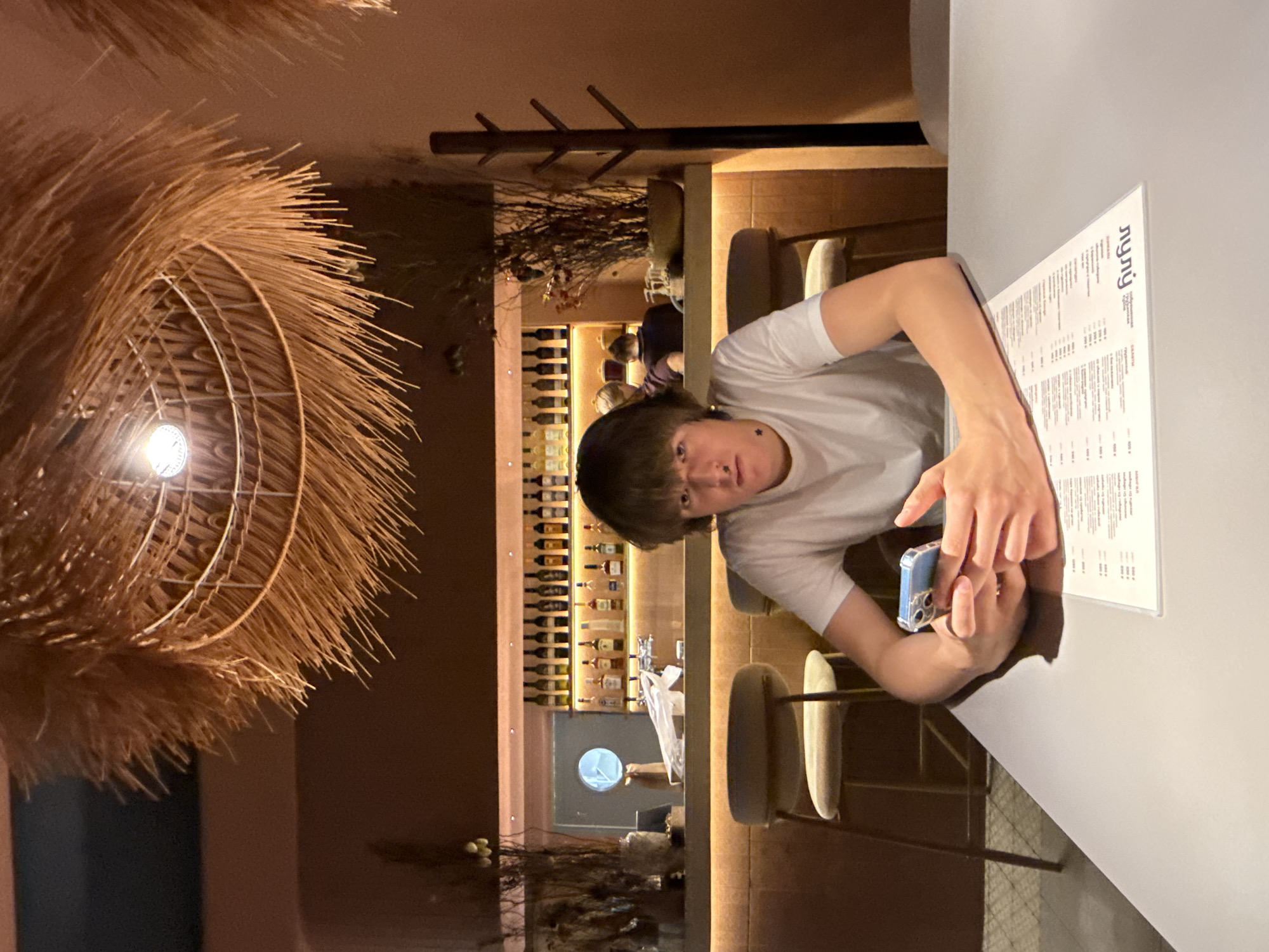 A young person with dark hair is seated at a table in a restaurant, holding a smartphone. The person is wearing a white t-shirt and has a nose ring. The table has a menu on it. Behind the person is a bar area with bottles on shelves. In the background, other patrons are visible. The restaurant has a modern design with a thatched light fixture hanging from the ceiling. The lighting is dim and warm. The image appears to have been taken at night. The location in the city of Chaikovsky, Russia is not discernible from the image.