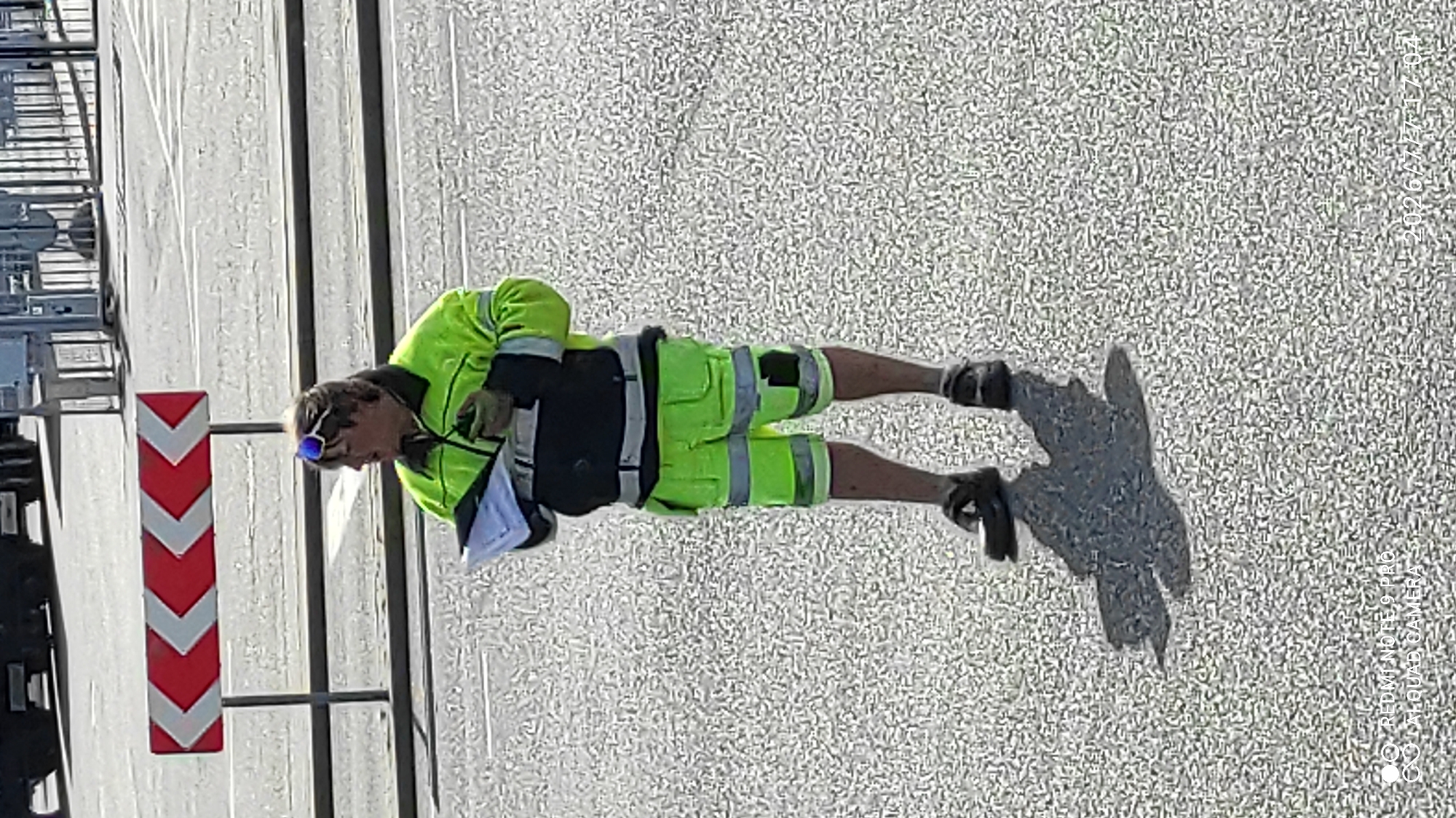 A person wearing a bright yellow high-visibility jacket and matching shorts with reflective stripes stands on a paved surface, holding a clipboard and a pen. They are also wearing dark shoes and sunglasses on their head. The background shows a red and white chevron road sign, indicating a hazard or direction. In the further background, there are structures that appear to be part of a port or industrial area, with fences and buildings. The ground is a speckled asphalt or similar pavement. The lighting suggests it is daytime. The watermark in the bottom left corner indicates the image was taken with a Redmi Note 9 Pro with a Quad Camera, and the bottom right corner shows a date and time: 2026/7/7 17:04. The setting is consistent with an industrial area or port, and considering the provided watermark includes "Tallinn, Estonia", it is likely this location.