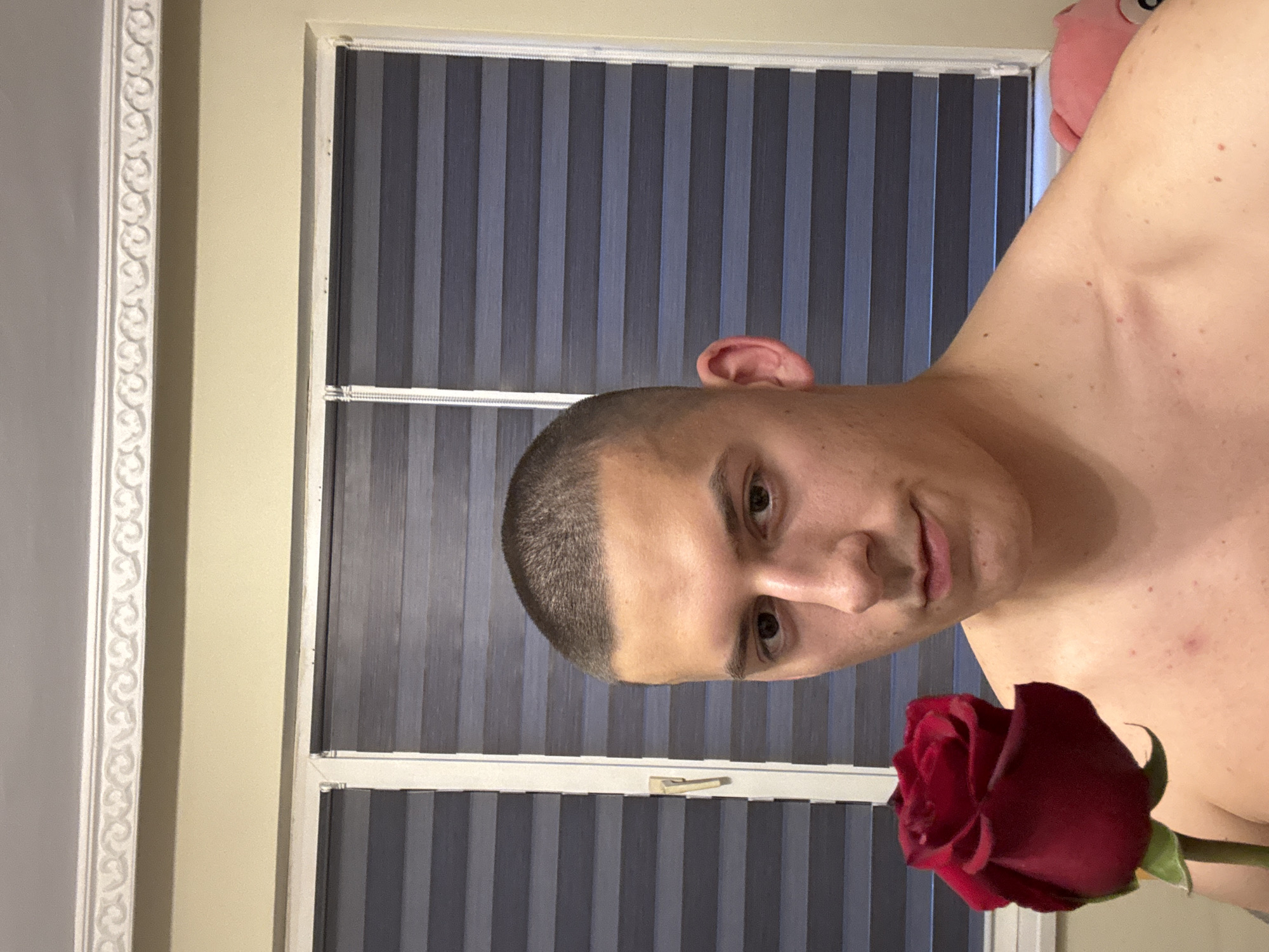 This media file, captured in Burgas, Bulgaria, presents a portrait of a young man holding a red rose. The man has short, dark, buzzed hair and fair skin, with visible moles or freckles across his chest and shoulder. He appears to be shirtless. He looks directly at the camera with a calm, slightly smiling expression.
In the foreground, a deep red rose with a green stem is held, partially obscuring the man's right shoulder and chest. The background features a window fitted with vertical blinds, displaying alternating stripes of dark and lighter blue-grey. A white window frame encloses the blinds. To the left of the window, a plain light beige wall is visible, bordered on its far left by decorative white molding or trim. In the upper right corner, behind the man's shoulder, a small pink object, resembling a plush toy, is faintly seen. The indoor setting with drawn blinds makes the specific time of day indeterminate. No text is visible in the image.
