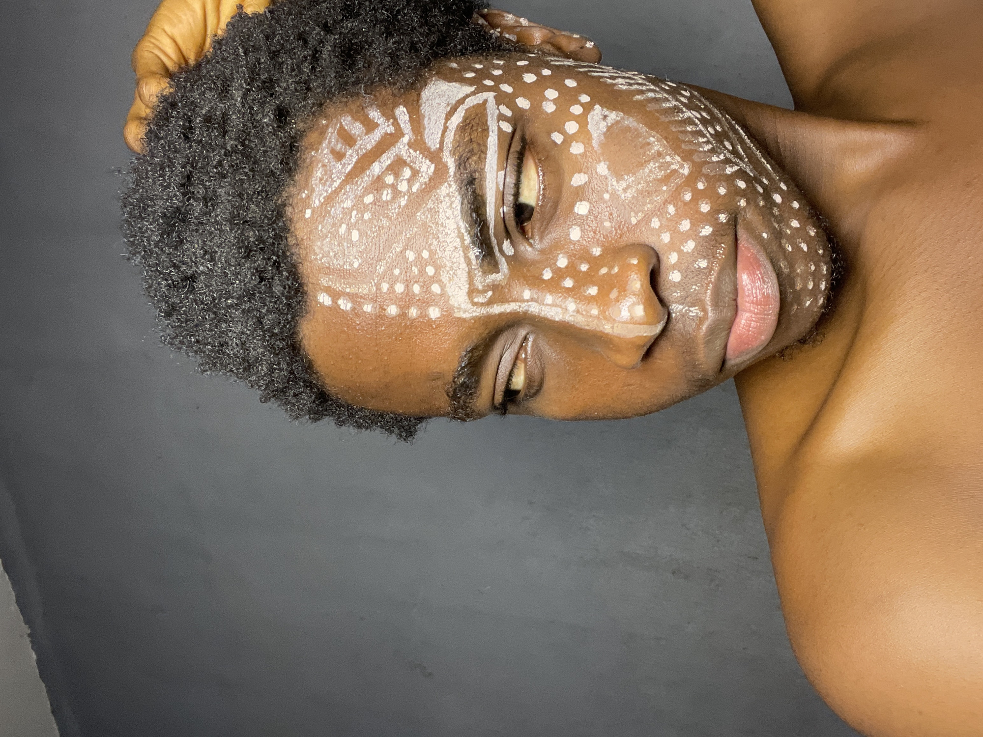 A person with dark skin and short, curly, dark hair is oriented at an angle against a dark gray background. The person's face is adorned with white paint in the form of dots and linear designs. The head is tilted towards the right, with the right shoulder visible. The individual is shirtless, revealing the upper chest and shoulder. No specific details indicate the city of Nkpor, Nigeria.