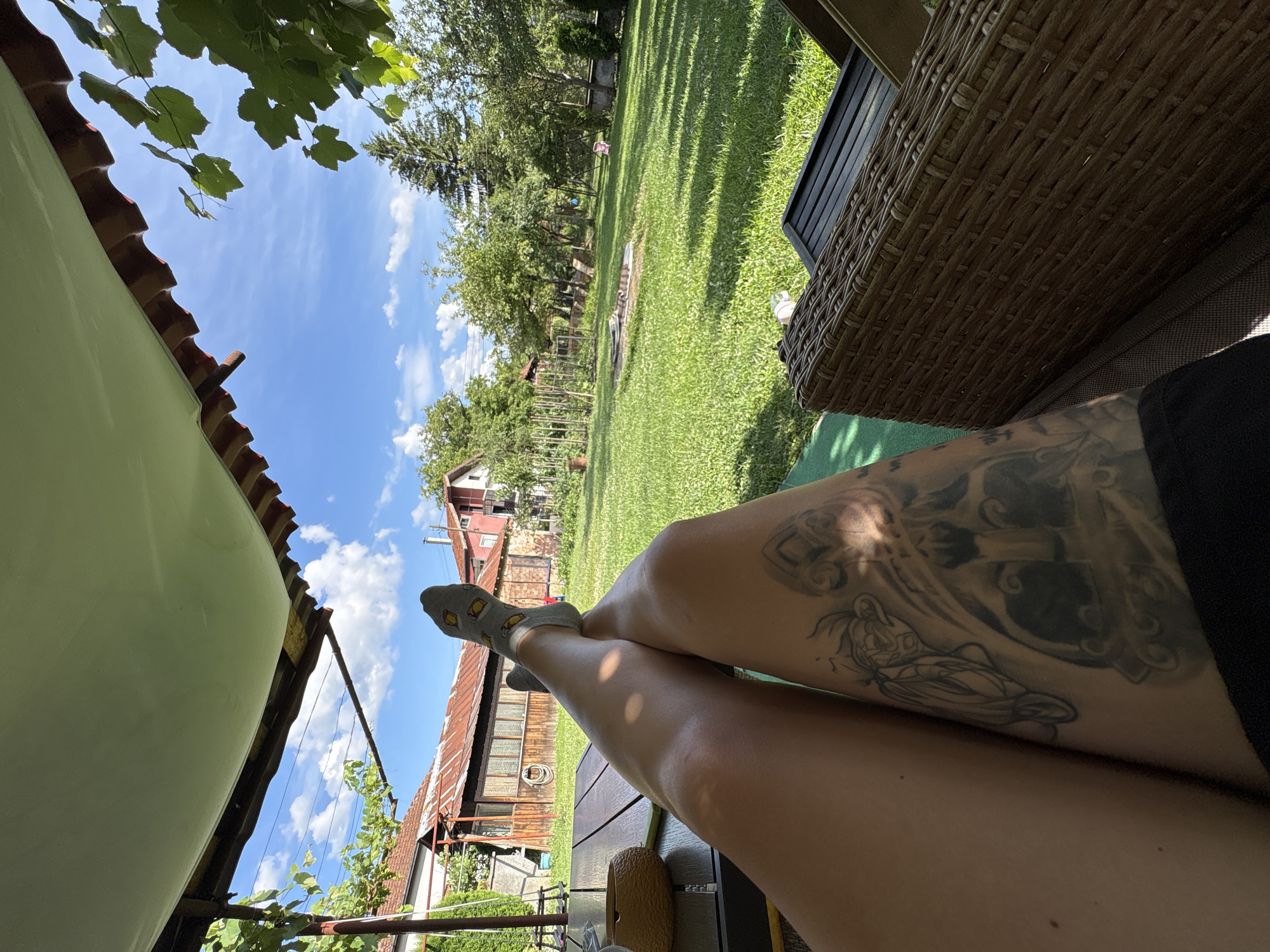 The image is a point-of-view shot from a person resting in a backyard in Oreshak, Bulgaria. The foreground features the person's legs, adorned with tattoos, resting on a brown wicker chair. One foot is visible, wearing a gray sock with a pattern. A wooden table is positioned to the left, with a woven brown object on it. The background reveals a lush green lawn extending into a garden area with trees and various structures, including houses with red roofs. The sky is blue with scattered white clouds. The overall scene depicts a relaxed outdoor setting during the daytime.