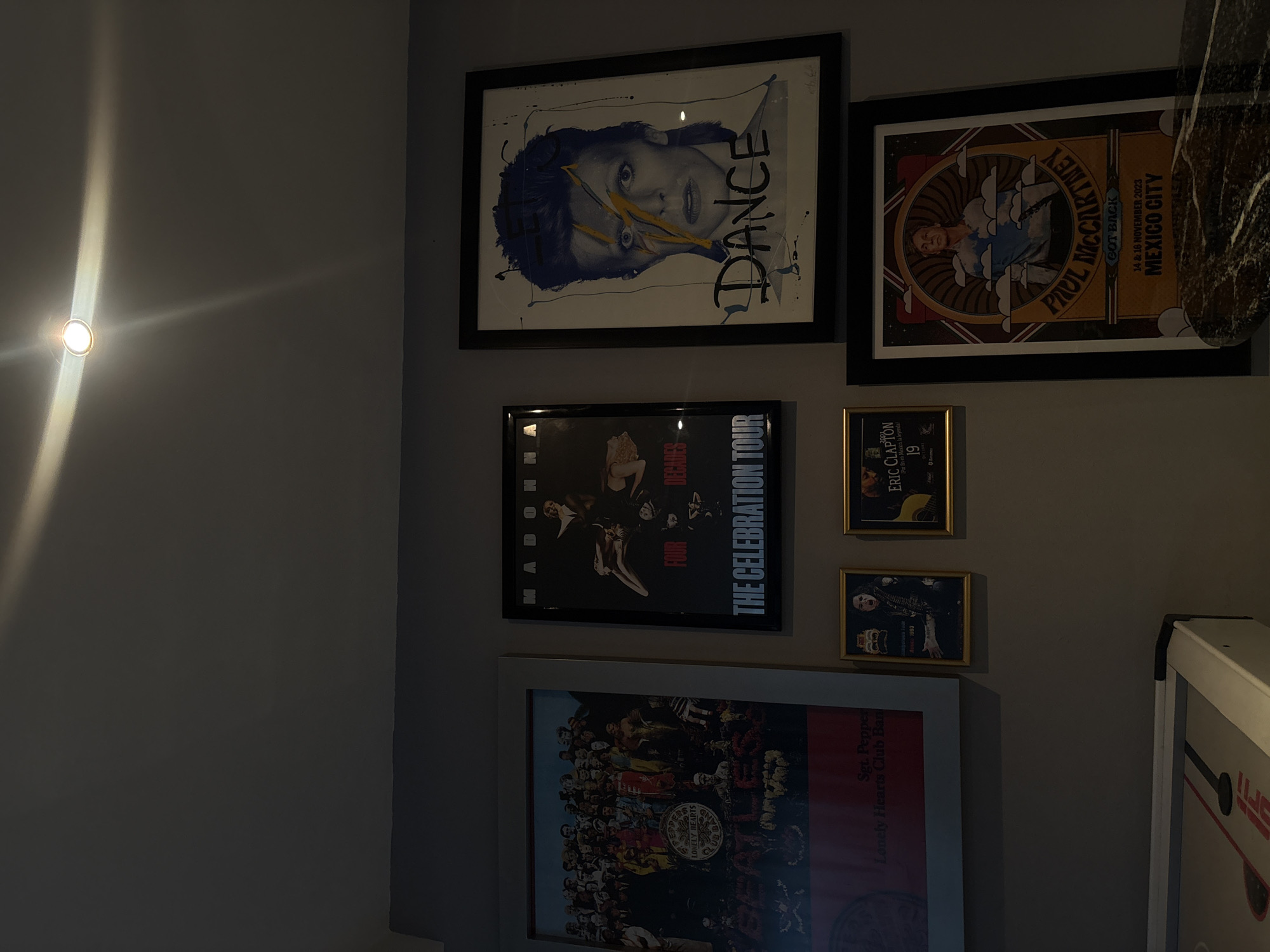 The image displays an indoor wall decorated with a collection of framed music posters and album art, illuminated by an artificial light source. The wall is painted a plain gray, and a bright, circular light fixture on the left casts a strong upward beam, creating a warm, elongated glow.
Several framed posters are visible in a dense arrangement. At the top left, a poster features a stylized face, possibly David Bowie, with blue and yellow accents and the prominent word "DANCE" written vertically. Adjacent to it, a poster advertises a "PAUL MCCARTNEY GOT BACK" concert in "MEXICO CITY" scheduled for "14 & 16 NOVEMBER 2023".
Below these, a poster for "MADONNA THE CELEBRATION TOUR" with the text "FOUR DECADES" is featured. To its right, two smaller framed pieces include an "ERIC CLAPTON" poster dated "1993", featuring the Spanish phrase "Por fin en México, la leyenda!". The bottom-most visible poster is the iconic album cover for The Beatles' "Sgt. Pepper's Lonely Hearts Club Band".
On the far right side of the image, a section of a white and red game table, possibly an air hockey table with "ESPN" branding, is partially visible. The overall lighting suggests an indoor setting, likely in the evening or night, creating a focused illumination on the displayed art. The posters and concert locations ("MEXICO CITY") align with the given context of San Bernardino Tlaxcalancingo, Mexico.