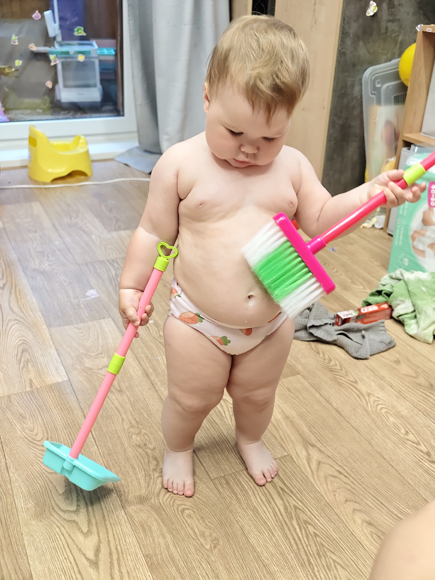A toddler with fair skin and blond hair is standing on a light-colored wooden floor in a room in Chaikovsky, Russia. The toddler is wearing patterned underwear and is bare-chested. The toddler is holding a child-sized pink and green broom in one hand and a child-sized pink and blue dustpan in the other. The background includes a window with a yellow object on the floor, a shelf, and various household items.