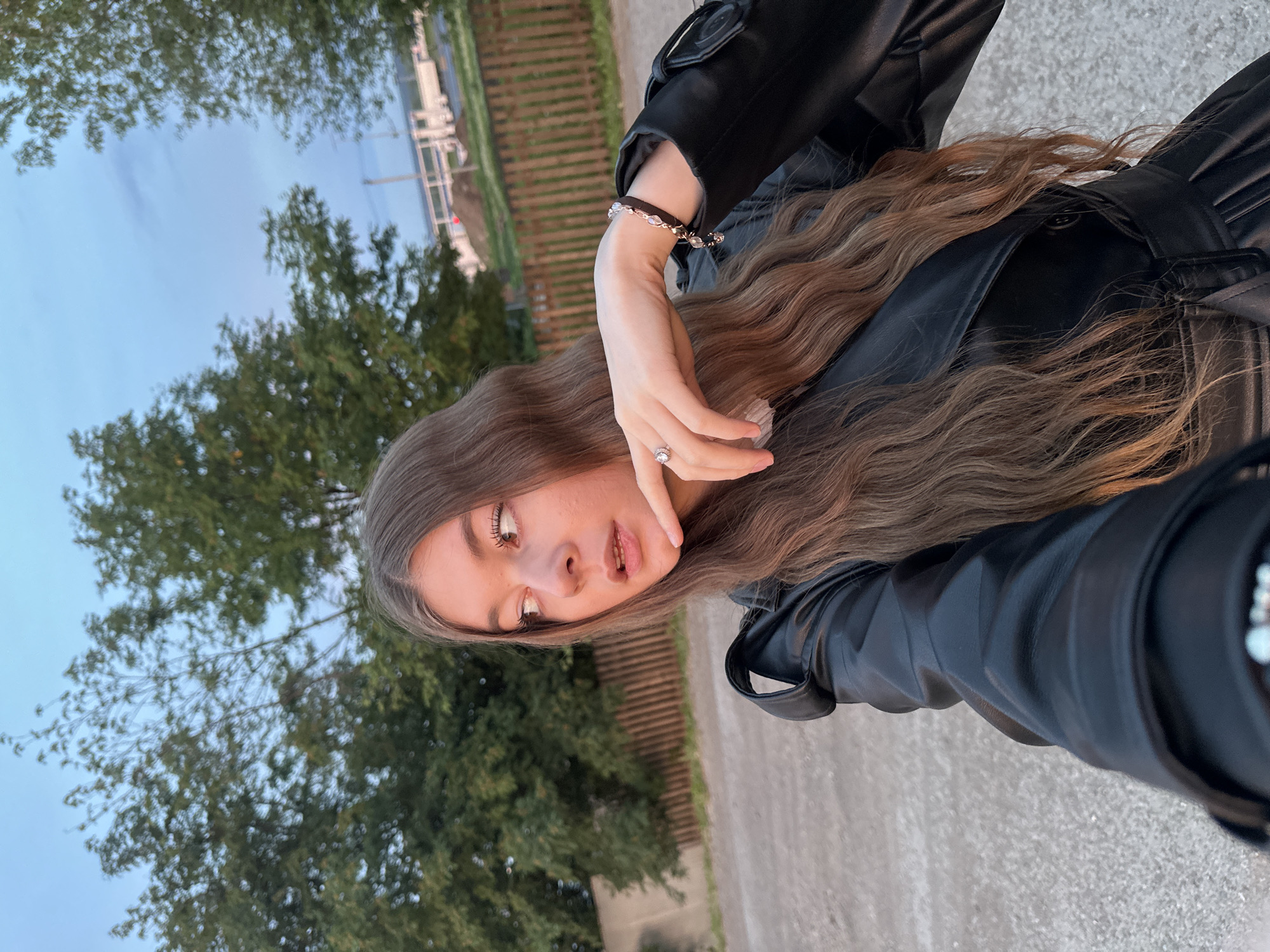 A young woman is seen in an outdoor setting, seemingly in or near Кузембетьево, Russia, given the prompt's instructions. She is wearing a black leather jacket. Her long, brown hair is wavy. She's wearing a bracelet on her left wrist. She also wears a ring on her left hand. The background consists of trees, a building in the distance, and what appears to be a paved surface. The sky appears to be transitioning in color, potentially indicating dawn or dusk.
