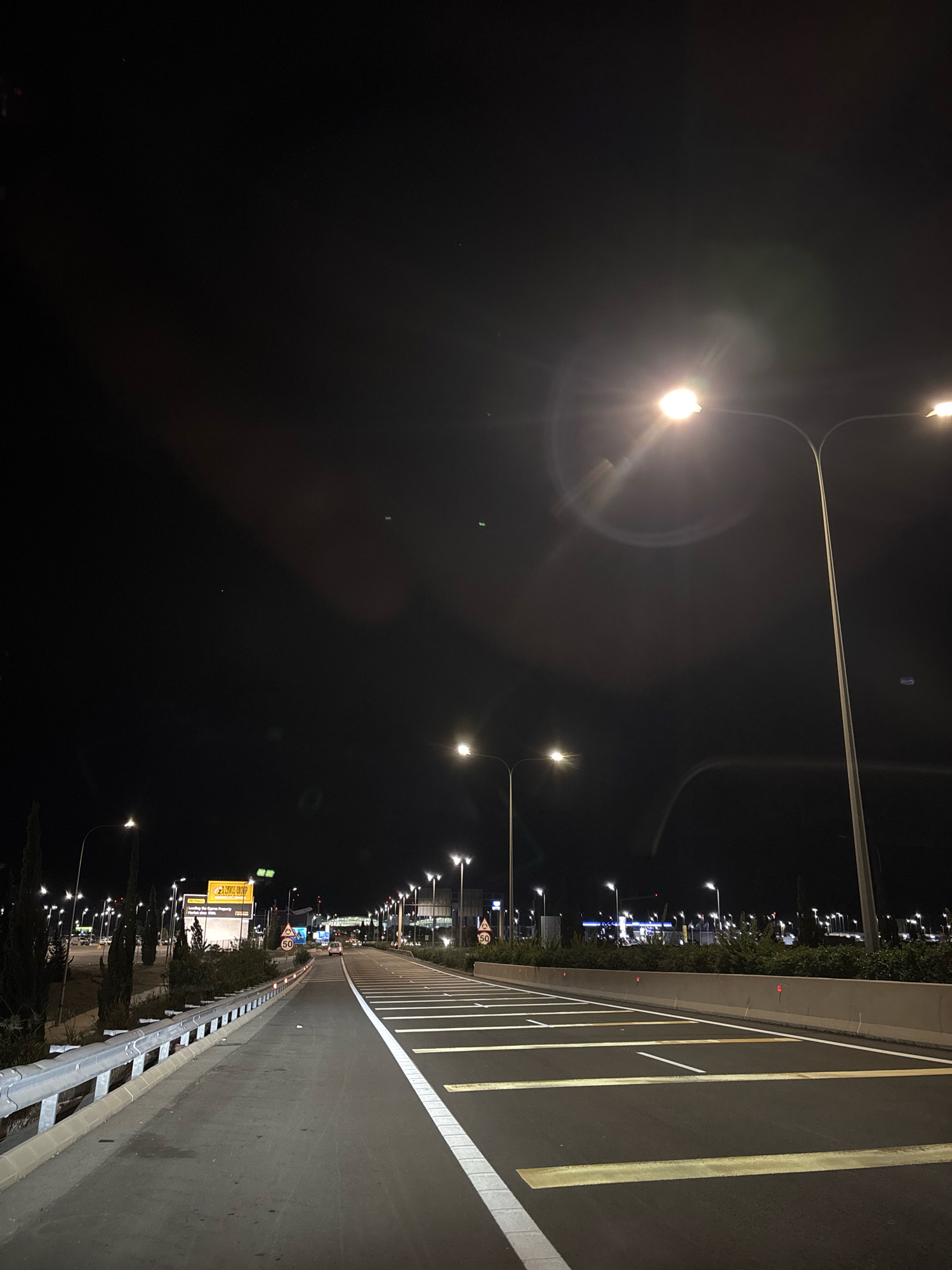 An aerial night shot shows a well-lit airport road in Dromolaxia - Meneou Municipality, Cyprus. The road has multiple lanes with clear white and yellow markings, indicating it is a primary thoroughfare. Streetlights line both sides of the road, casting a bright glow and creating lens flares due to the dark sky. On the left side, there is a highway guardrail and some vegetation, including cypress trees. A large yellow billboard advertising "LEWIS GROUP" is visible on the left, with a speed limit sign of "50" displayed on the right. In the distance, airport terminal buildings and more lighting suggest an active airport environment. The sky is dark, with a few faint lights or stars. There are no people or vehicles clearly visible in the immediate foreground, but faint car lights are seen in the distance. The overall scene depicts a modern, organized infrastructure at night.