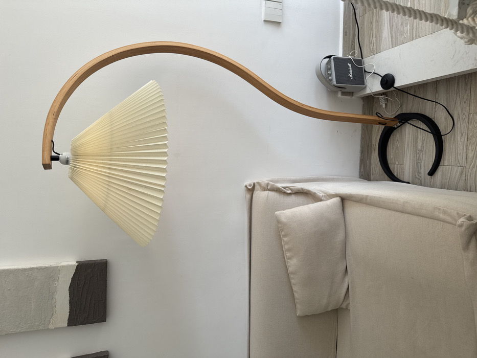 The image displays an interior room characterized by light-colored walls and a matching floor. On the left, a beige, slipcovered sofa with one rectangular beige cushion is partially visible. Above the sofa, two vertical rectangular artworks with abstract, textured grey, white, and dark brown segments are mounted on the wall. To the right of the sofa, a tall floor lamp features a curved, light-brown wooden stand terminating in a pleated, conical, cream-colored lampshade. The lamp's base is a black crescent shape. Adjacent to the lamp's base and against the wall, a grey Marshall brand portable speaker is positioned on the floor. Black electrical cords extend from both the lamp (including a circular foot switch) and the speaker, leading towards a white multi-slot electrical outlet visible on the wall behind the speaker. The floor is light-colored, possibly wood or laminate, with a low, white rectangular platform running along the bottom right edge. A white braided rope is partially visible on the far right. No individuals are present in the scene.