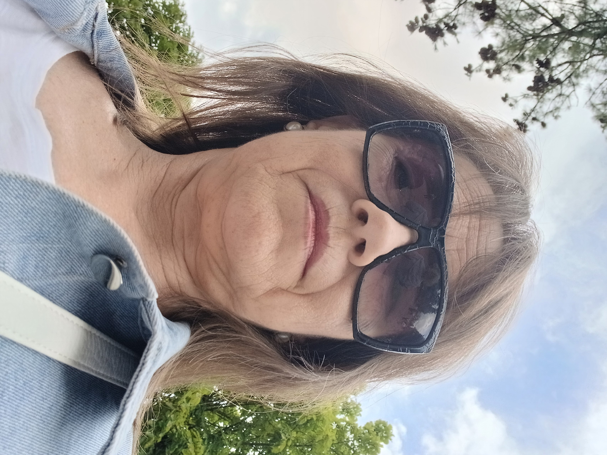 A medium shot of a woman wearing large, dark sunglasses and a denim jacket, taken from a low angle. She has light brown hair and appears to be outdoors, with trees and a cloudy sky in the background. The image does not provide any information that would allow for its location to be determined in Truskavets, Ukraine or any other specific location.
