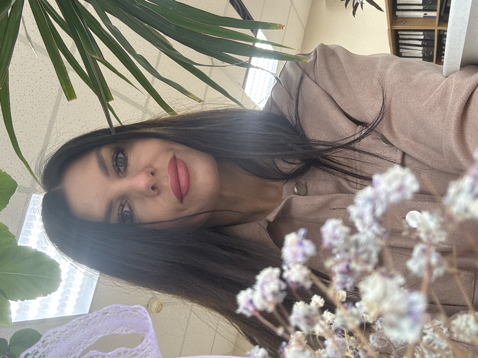 The media file, an image rotated 90 degrees counter-clockwise, presents a close-up portrait of a woman captured from a low angle. She has long, dark brown hair that falls over her shoulders, striking light-colored (likely blue or grey) eyes, and full lips accentuated by pink lipstick. Her gaze is directed slightly upwards, and her expression is serene. She is dressed in a light brown or beige jacket, possibly made of suede or a similar soft-textured fabric, with a prominent button visible on the front.
In the left foreground, large, vibrant green leaves of an indoor plant partially obscure the view, adding a touch of nature. On the right, a cluster of out-of-focus white and pale purple dried flowers (resembling baby's breath) contributes to the soft foreground elements.
The background reveals an indoor setting, likely an office or study. A textured white ceiling with bright overhead fluorescent light fixtures is visible, suggesting artificial illumination. In the upper right corner, a wooden bookshelf filled with binders and files provides a clear indication of a professional or academic environment. The composition, with the subject looking up and the ceiling visible, suggests a selfie taken from a low vantage point. No discernible text is present in the image. The scene is set indoors, potentially in Chaikovsky, Russia.