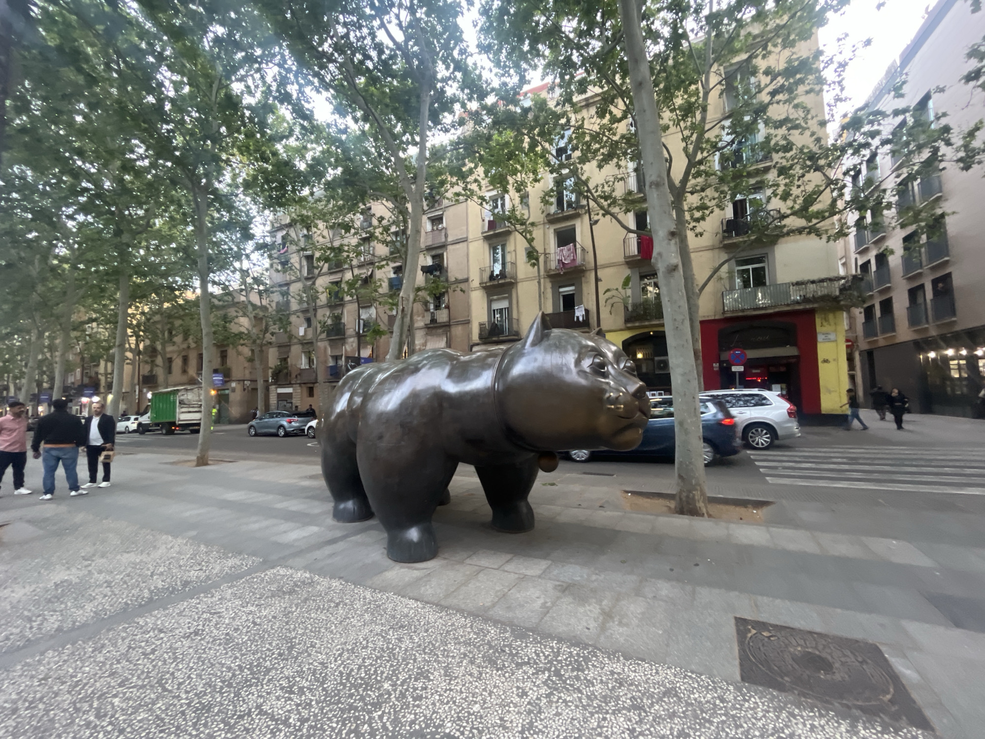The image depicts a bustling urban street scene in Barcelona, Spain, characterized by a large, dark bronze statue of a stout, hippopotamus-like animal, likely a work by Fernando Botero, positioned prominently on the sidewalk in the foreground.
The street is a wide boulevard, lined with numerous mature trees featuring green foliage that forms a partial canopy overhead. Multi-story buildings with light-colored facades and balconies, typical of European architecture, flank the street. Some balconies display clothes drying, adding a touch of everyday life.
On the left side of the sidewalk, three men are visible: one in a pink shirt, another in a dark top and blue jeans, and a third in a white jacket over a dark shirt. They appear to be conversing. In the background, various vehicles, including a green truck, a white SUV, a grey sedan, and a blue car, are present on the street, indicating active traffic. Several blurred figures of pedestrians are also seen walking across the street or on the opposite sidewalk.
A red storefront entrance, possibly belonging to a restaurant or bar, is visible in the mid-ground, with the faint text "La Taverna" discernable on its sign. A decorative manhole cover is visible on the paved sidewalk in the foreground.
The lighting suggests it is daytime, possibly late afternoon or early evening, with a diffused light quality and mild shadows. The weather appears clear and pleasant. The scene captures a typical, lively city environment.