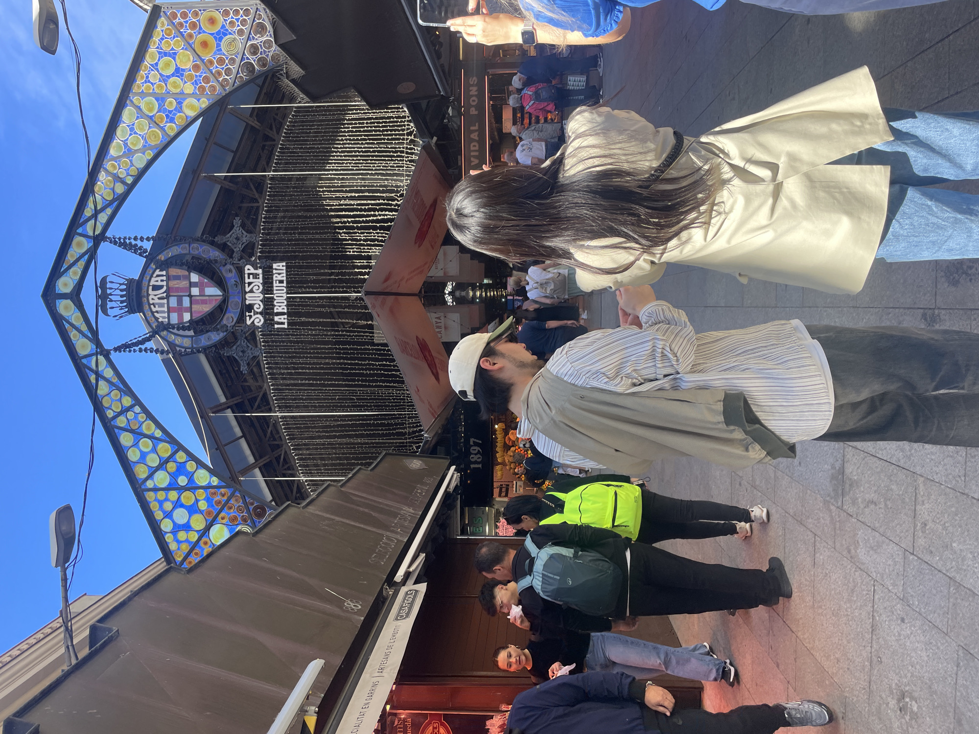 The image captures a street scene in front of the Mercat de Sant Josep de la Boqueria, Barcelona. A brightly decorated entrance with colored glass and the market's name arches over the entrance. Several people are in the frame, facing in various directions and engaged in different activities. A woman with long dark hair is wearing a beige trench coat and blue jeans, partially blocking the view of the market entrance. Next to her is a person in a striped shirt, cap, and khaki-colored jacket. Below them are other people, including a person with a bright green backpack and a group seemingly eating or handling food. The environment includes various storefronts and signage, indicating shops and restaurants within the market area. The cobblestone ground and architecture of the market are visible.