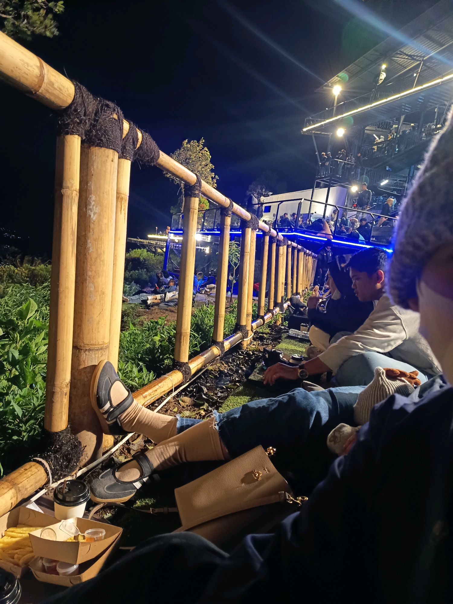 In Keditan, Indonesia, a group of individuals is gathered outdoors at night. They are seated on the ground in front of a bamboo fence. Some people are looking towards a multi-story structure with bright lights and numerous people on its platforms. Food items, including french fries and drinks in paper cups, are visible on the ground. The environment suggests a casual outdoor gathering or event.