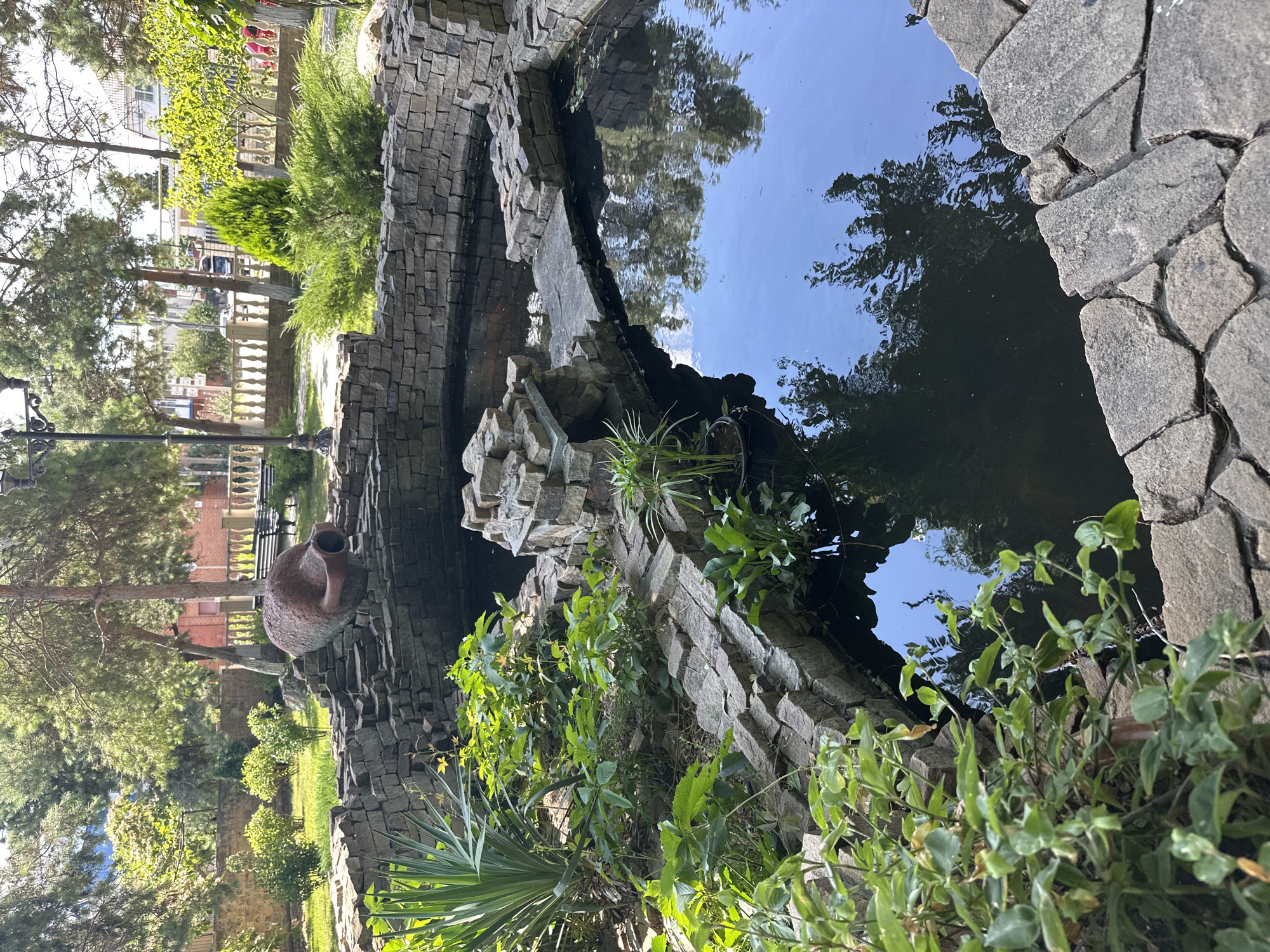 The image depicts a decorative pond or water feature constructed with rough-hewn stones. A large, reddish-brown amphora lies on its side, partially embedded in the stone structure at the top of the pond. Lush green vegetation grows around the edges of the pond, with some plants appearing to be aquatic. The water surface reflects the surrounding trees and sky. In the background, there is a paved area, a lamppost, a white balustrade, and buildings, suggesting an urban or park-like setting. The scene is located in the city of Taman, Russia.
