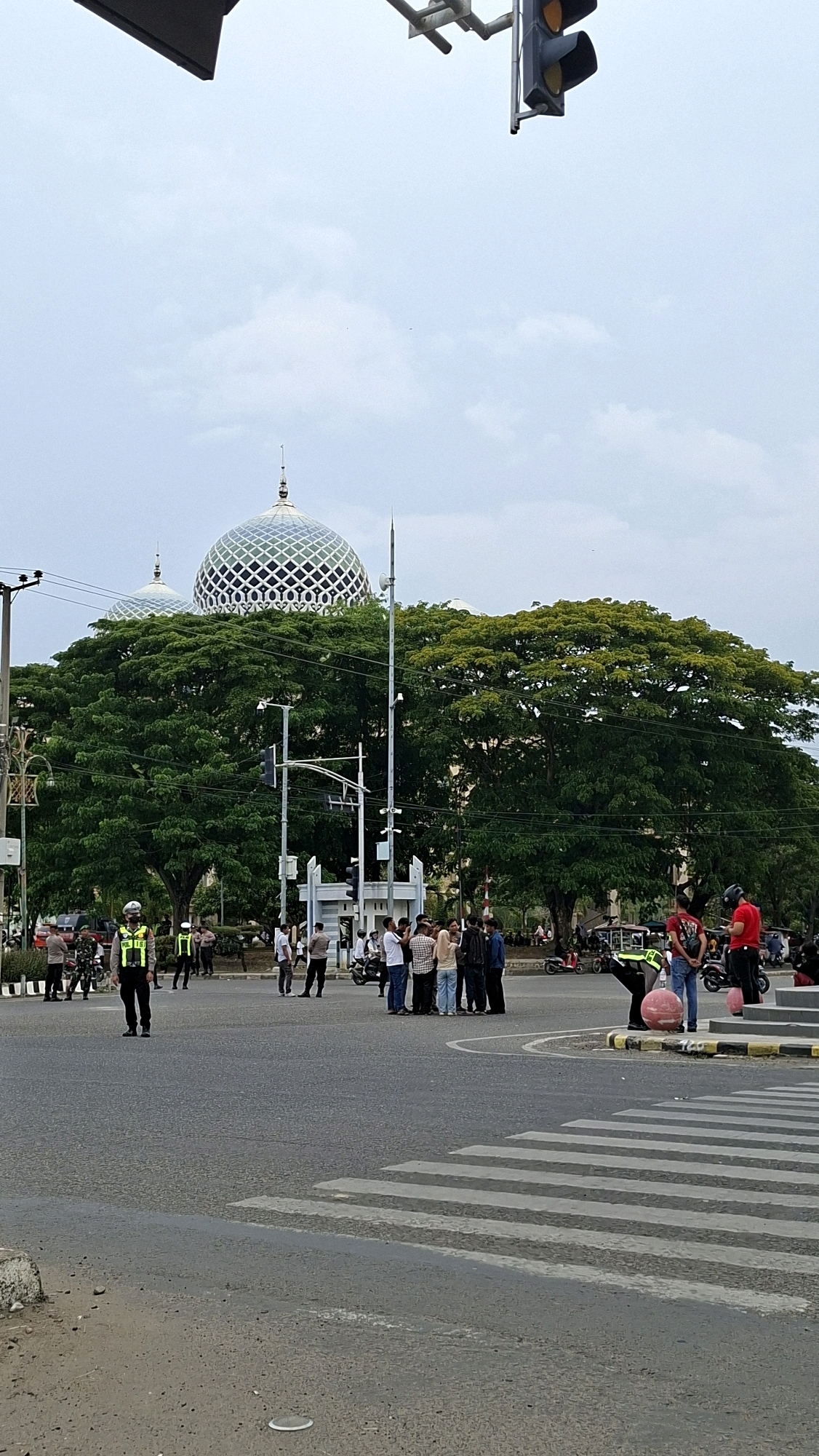 The image depicts a busy intersection in Lhokseumawe, Indonesia, with the distinctive domes of the Islamic Center Lhokseumawe prominently featured in the background. The sky is overcast. In the foreground, there is a crosswalk. Several individuals are present, including police officers in uniform directing traffic and a group of civilians gathered. Some are on foot, while others are near motorcycles. The atmosphere appears to be one of controlled activity, possibly a traffic management situation or a gathering. Large, mature trees with dense green foliage surround the area, providing a natural element to the urban landscape. Traffic signals and streetlights are visible, indicating the presence of active infrastructure.