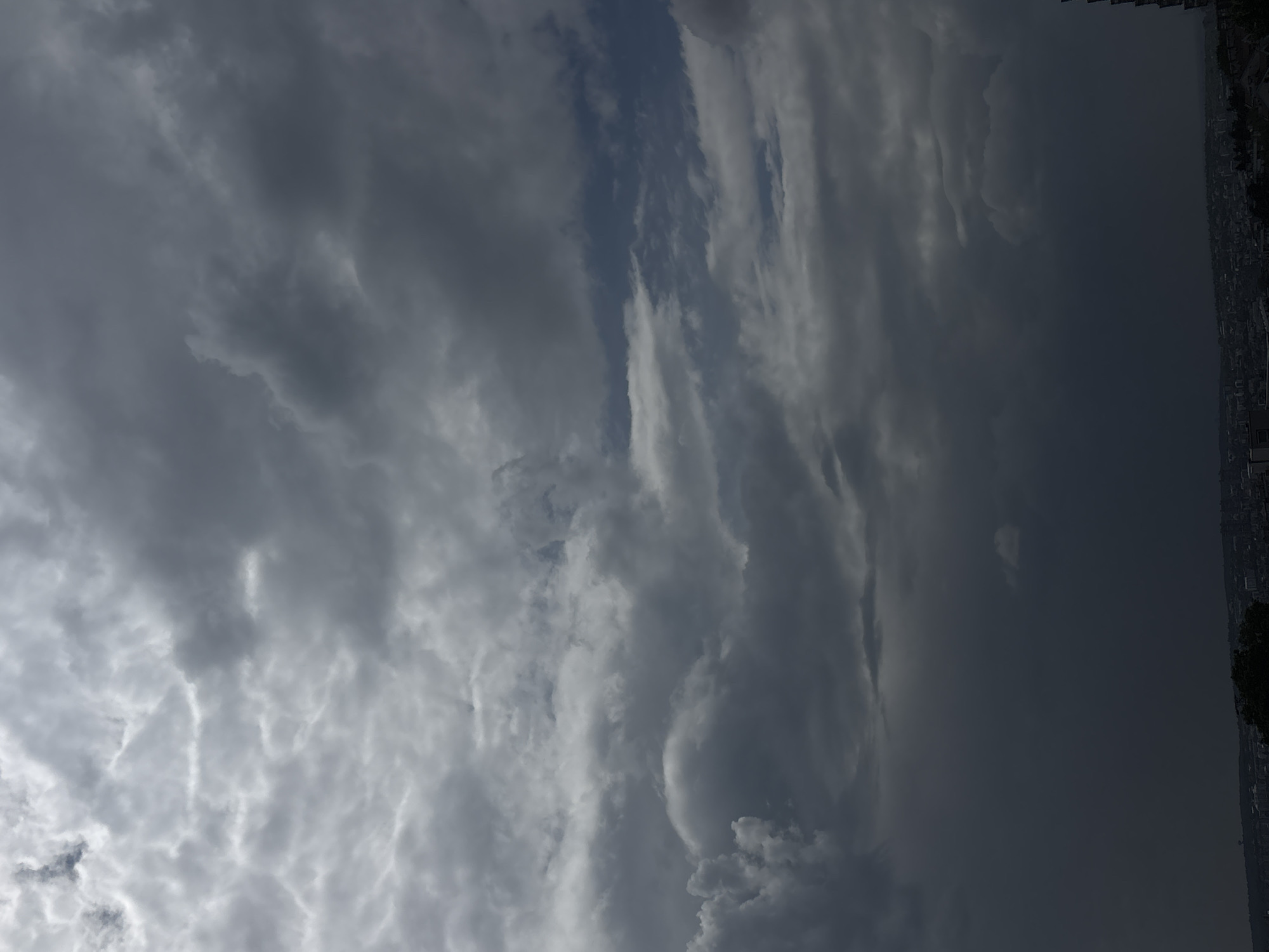 The image depicts a cloudy sky over Belgrade, Serbia. To the right side of the image, a cityscape can be seen. The sky is filled with various cloud formations, ranging from thin, wispy clouds to dark, ominous ones. The lighting suggests a potentially stormy weather pattern. The focus is predominantly on the sky, with the city serving as a background element.