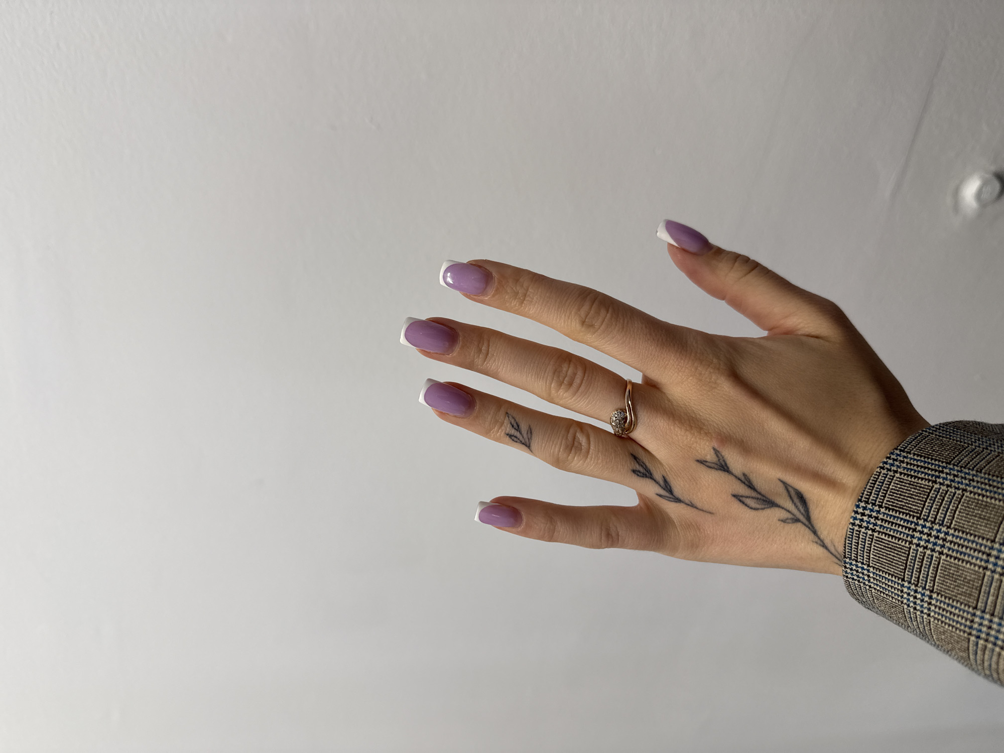 A close-up, eye-level shot focuses on a person's left hand, adorned with a delicate twig tattoo that extends from the wrist up the back of the hand. The hand is lightly tanned and features manicured nails painted with a lavender hue and white tips, a French manicure style. A thin, rose gold ring with a small, sparkling stone is worn on the ring finger. The person is wearing a textured, grey and blue plaid blazer. The hand is positioned against a plain, textured white wall. The setting is indoors, with no specific identifiable landmarks of Łódź, Poland visible. The lighting is even, suggesting a well-lit room or studio environment.