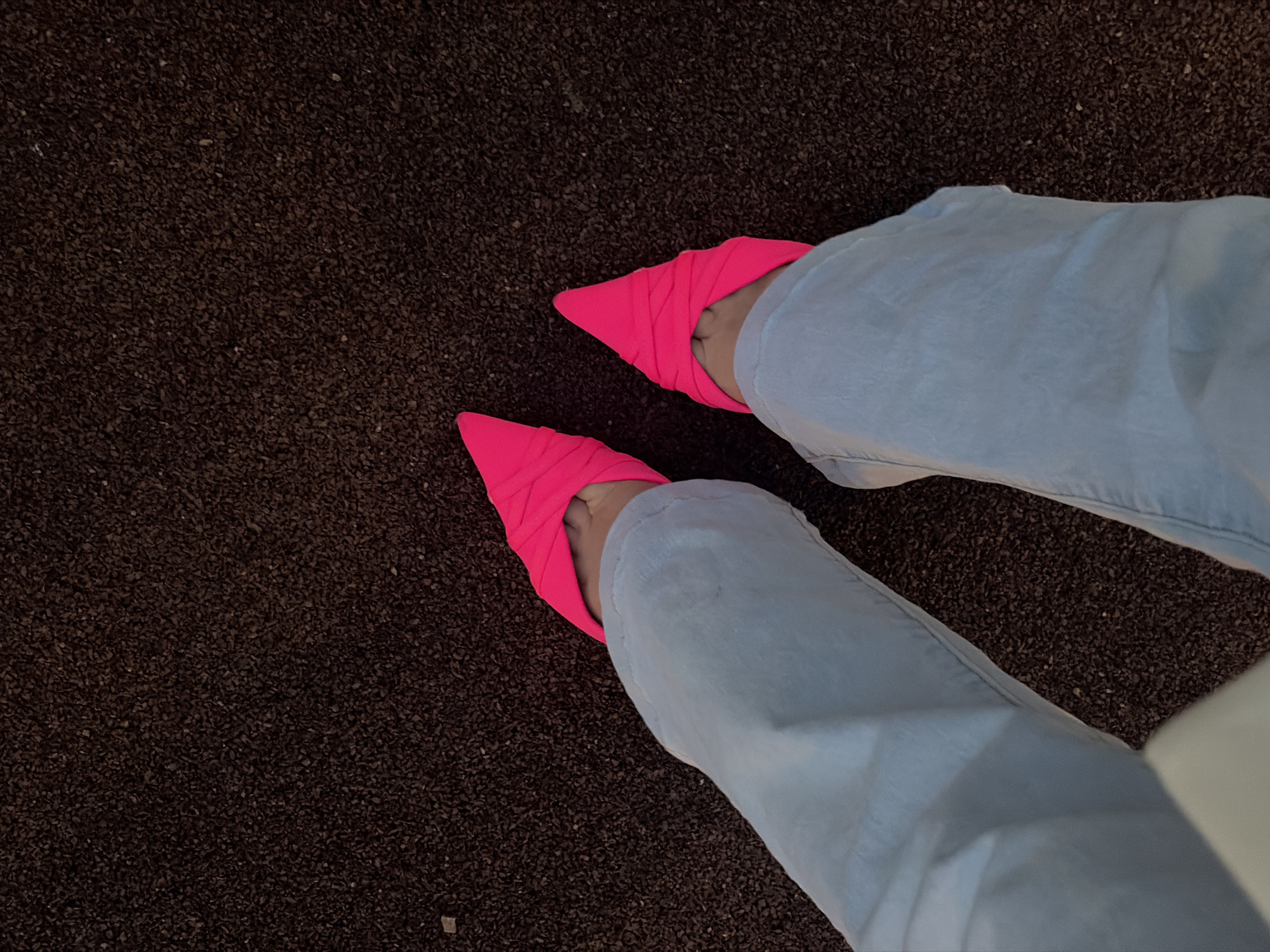 Two human feet, adorned with bright pink pointed-toe heels, are positioned on a dark, granular surface. The feet are clad in light blue, creased trousers. The background consists entirely of the dark, textured material. The scene appears to be outdoors, possibly on a paved area with a rubberized or gravelly finish, common in public spaces such as parks or playgrounds. There is no visible indication of the city of Saint Petersburg, Russia.