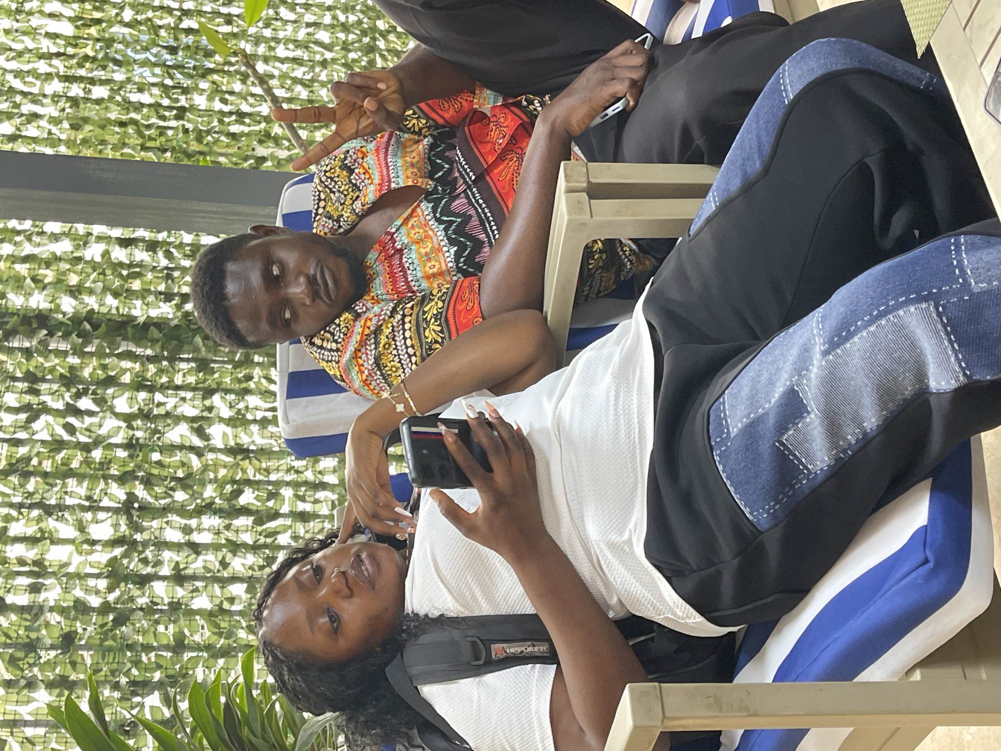 This image features two individuals, a man and a woman, relaxing in an outdoor or semi-outdoor setting.
The man, with a dark complexion and short dark hair, is wearing a vibrant, multi-colored patterned shirt (featuring red, yellow, black, and white hues) and dark trousers. He is making a peace sign with his right hand and holding a smartphone in his left hand. He looks towards the camera with a calm expression.
The woman, also with a dark complexion and long, curly dark hair, is reclining next to him. She is dressed in a white textured top and black trousers that have distinct blue denim patches on the sides. A black strap, likely from a backpack or harness, crosses her chest, bearing the visible text "HIPPOWER". She holds a black smartphone with a case in her hands, showcasing her long, decorated nails. She is also looking towards the camera with a relaxed demeanor.
Both individuals are seated or lounging on chairs equipped with blue and white striped cushions. The background consists of a green foliage wall or canopy with a grid-like pattern, suggesting a shaded and pleasant outdoor environment. The lighting indicates it is daytime. The overall scene conveys a relaxed and casual mood. The location is specified as Abuja, Nigeria.