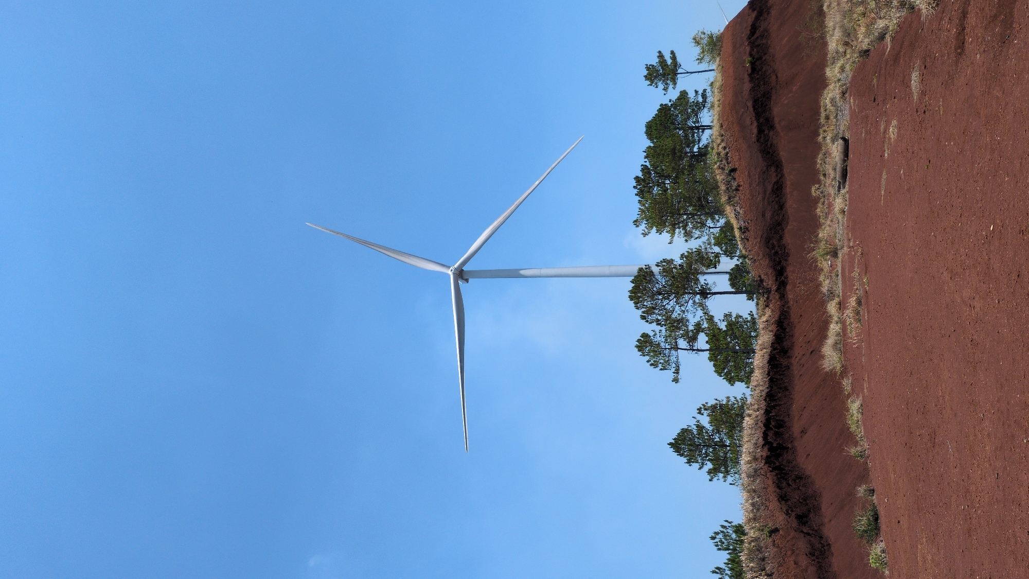 The image shows a wind turbine set against a clear blue sky. The foreground features a red dirt embankment with sparse vegetation and trees. The location, Phát Chi, Vietnam, is not apparent from the visual content of the image. There are no people visible.