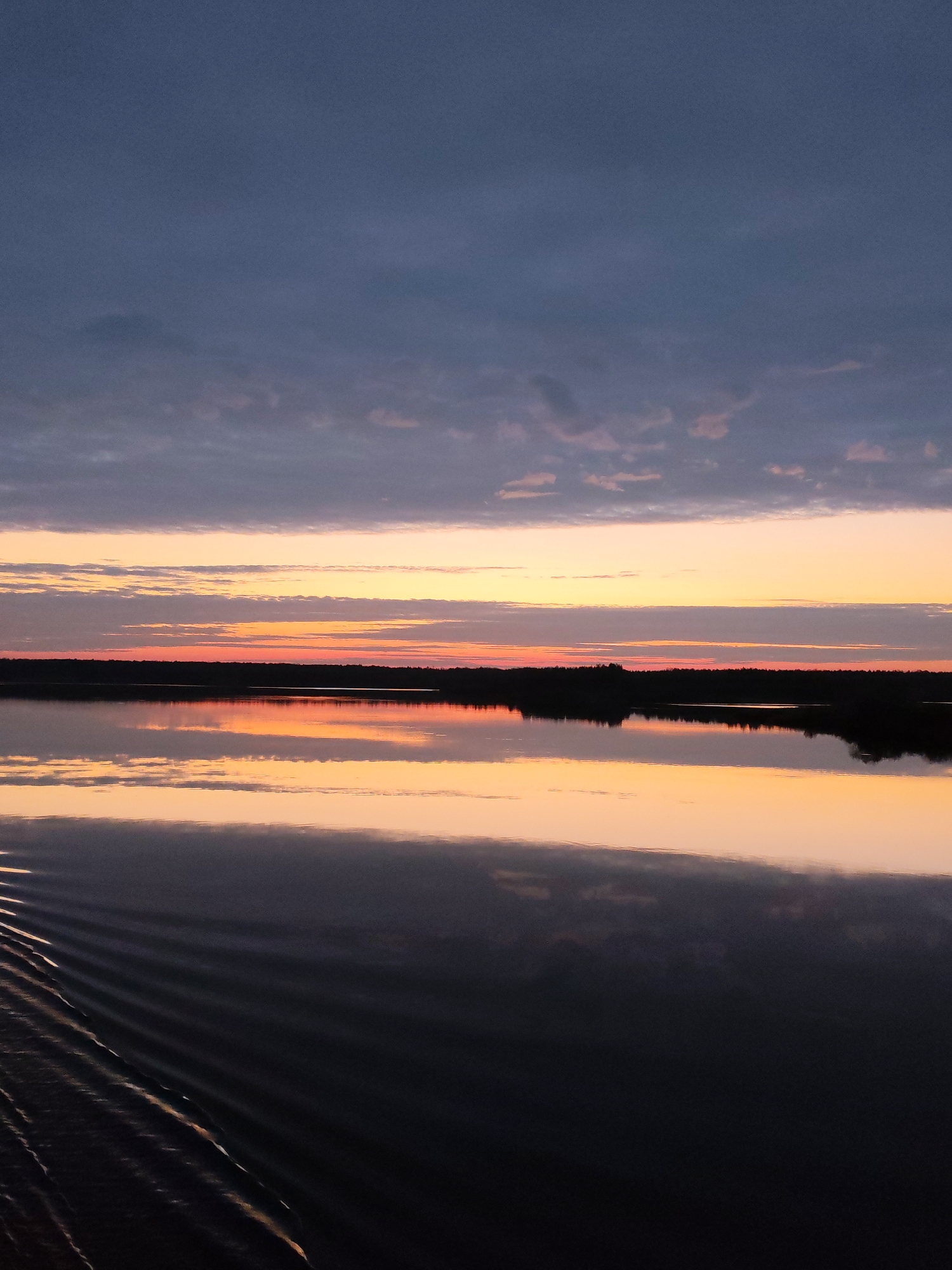 The image captures a serene landscape at dawn or dusk. The sky is a gradient of colors, transitioning from a deep blue at the top to bands of orange and pink near the horizon, with scattered clouds. Below the horizon lies a body of water, likely a lake or wide river, mirroring the colors of the sky, creating a symmetrical effect. A line of trees is present along the horizon, silhouetted against the colorful sky. In the foreground, there are ripples on the water surface, suggesting the presence of a gentle breeze or the movement of a boat. Based on the location, Даргун, Russia, the scene is likely a reflection of the surrounding natural landscape.