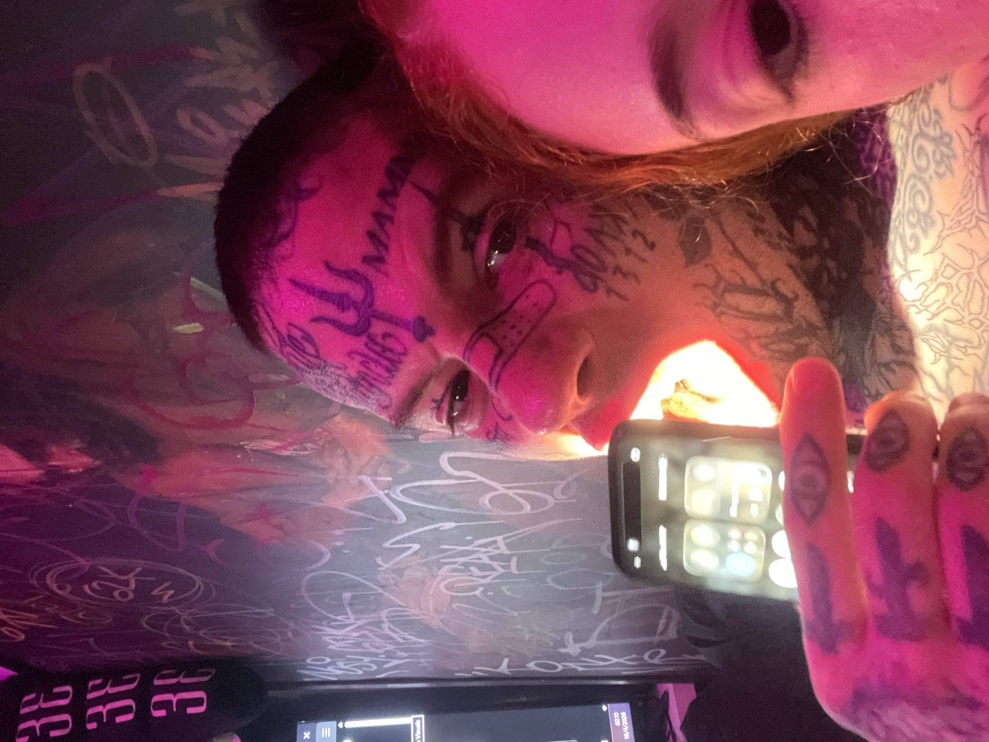 A photograph features two people, one holding a smartphone, under a pink-toned light source. The person in the foreground is partially visible, focusing on their face adorned with tattoos, including inscriptions and symbolic imagery. They have a band-aid on their nose. The smartphone in their hand displays a bright screen with app icons. The other person is only partially in the frame with their right eye visible. Visible in the background is the top of a desk covered with graffiti in a pale color. A piece of equipment is also visible in the bottom part of the image. The location and city of Buenos Aires, Argentina, cannot be determined from the information present in the photograph.