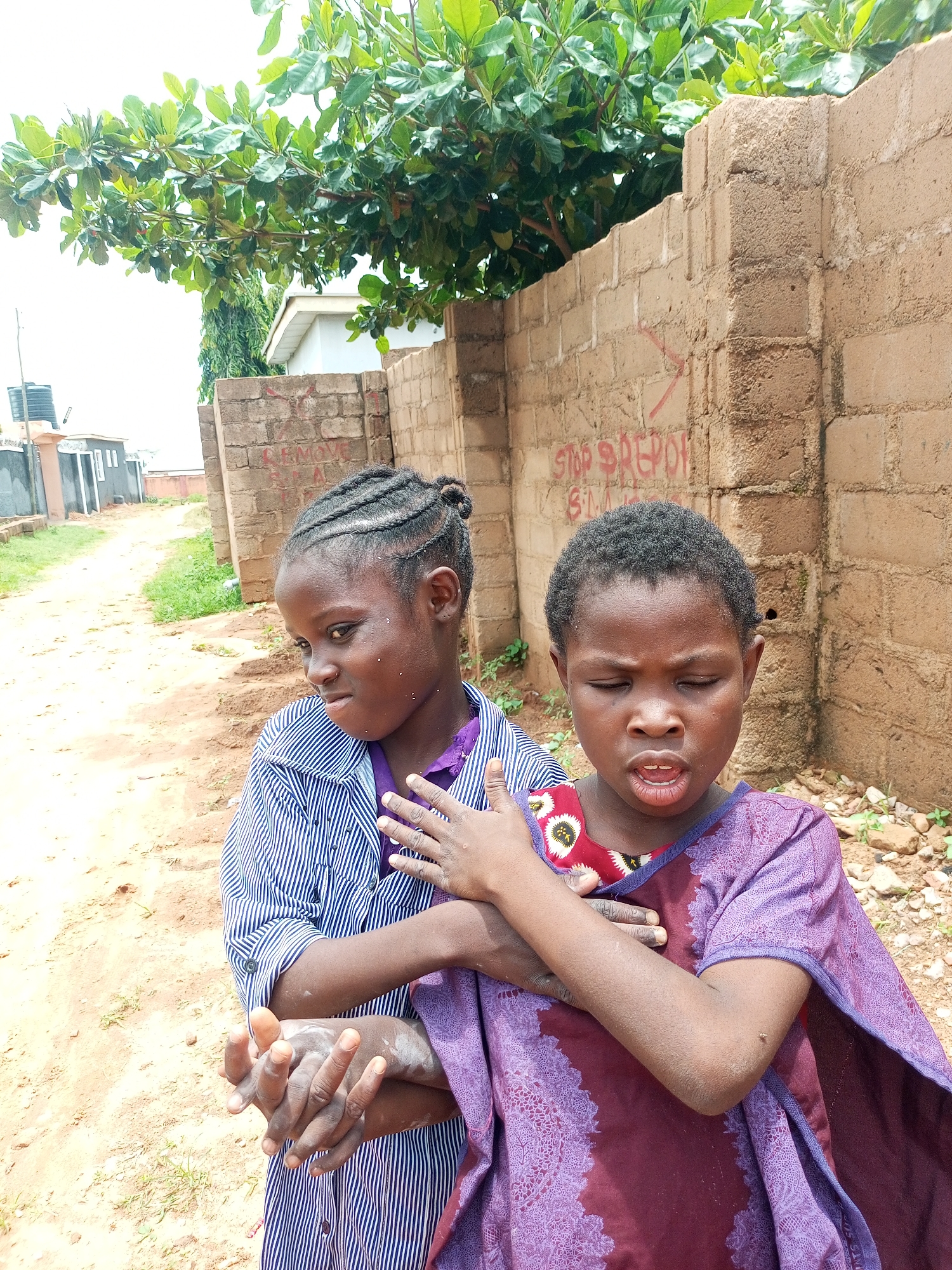 Two young girls, appearing to be of African descent, stand outdoors in what seems to be an urban environment. The girl on the left, facing right with her head turned slightly, has her hair braided into cornrows and pulled back into a bun. She is wearing a blue and white striped shirt over a purple garment. Her hands are clasped together in front of her. The girl on the right, facing forward with her eyes closed and mouth open as if speaking or singing, has short, dark hair. She is wearing a purple dress with a patterned red and white circular design on the chest area. Her arms are crossed, with her right arm over her left. Both girls have dirt on their skin and clothing.
The background consists of a rough, tan brick wall with visible graffiti. To the left of the wall, a dirt path leads into the distance, bordered by sparse green vegetation and some buildings. Overhead, lush green leaves from a tree branch extend into the frame. The overall lighting suggests it is daytime, possibly overcast. The scene is located in the city of Tempure, Nigeria.