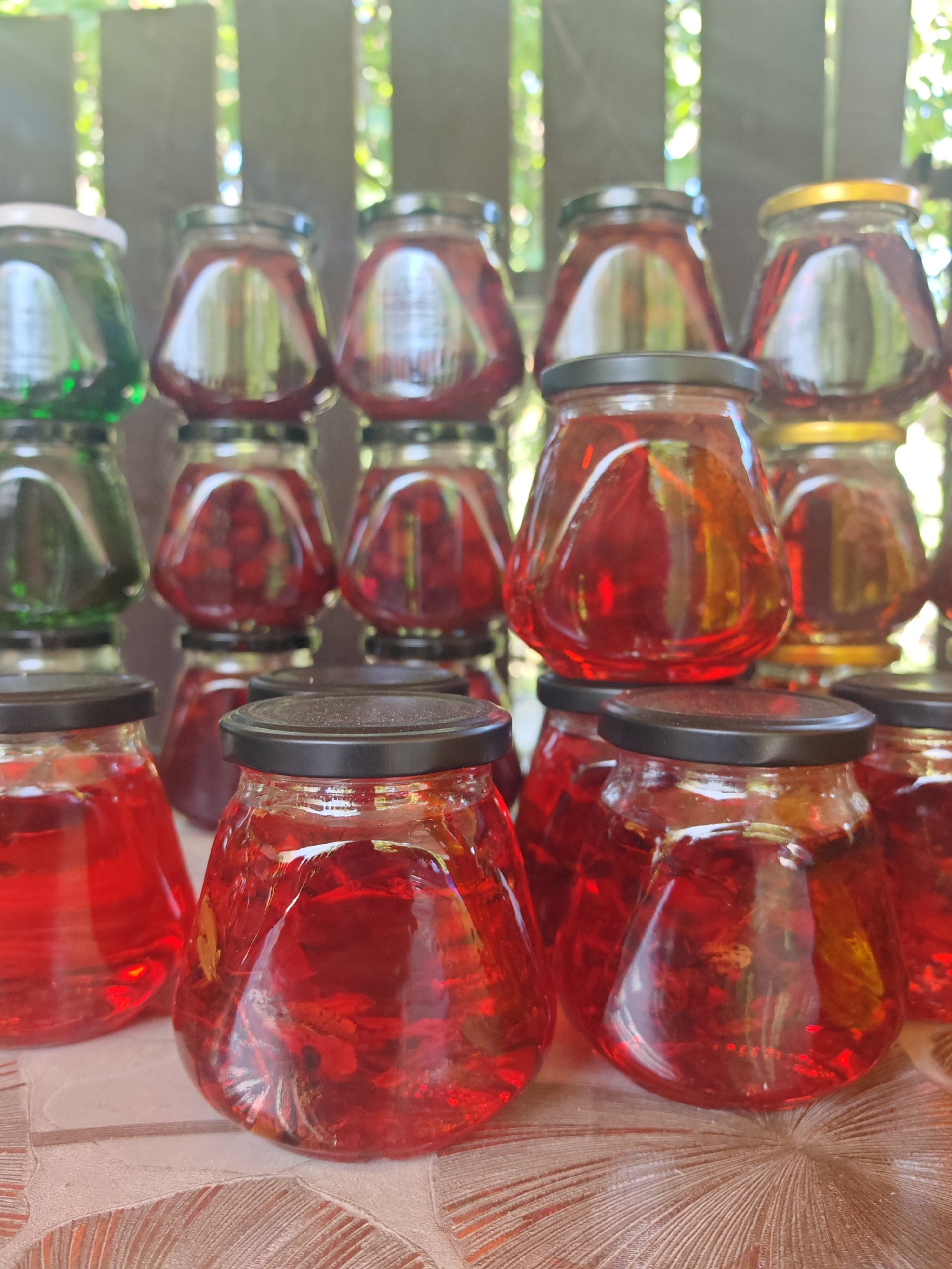 A collection of glass jars filled with red liquid, possibly jam or preserves, are arranged on a table. The jars are of various sizes and shapes, and most have black lids, with a few having gold or white lids. The background consists of a wooden fence and blurred green foliage, suggesting an outdoor setting. The scene appears to be located in Varvarovka, Russia.