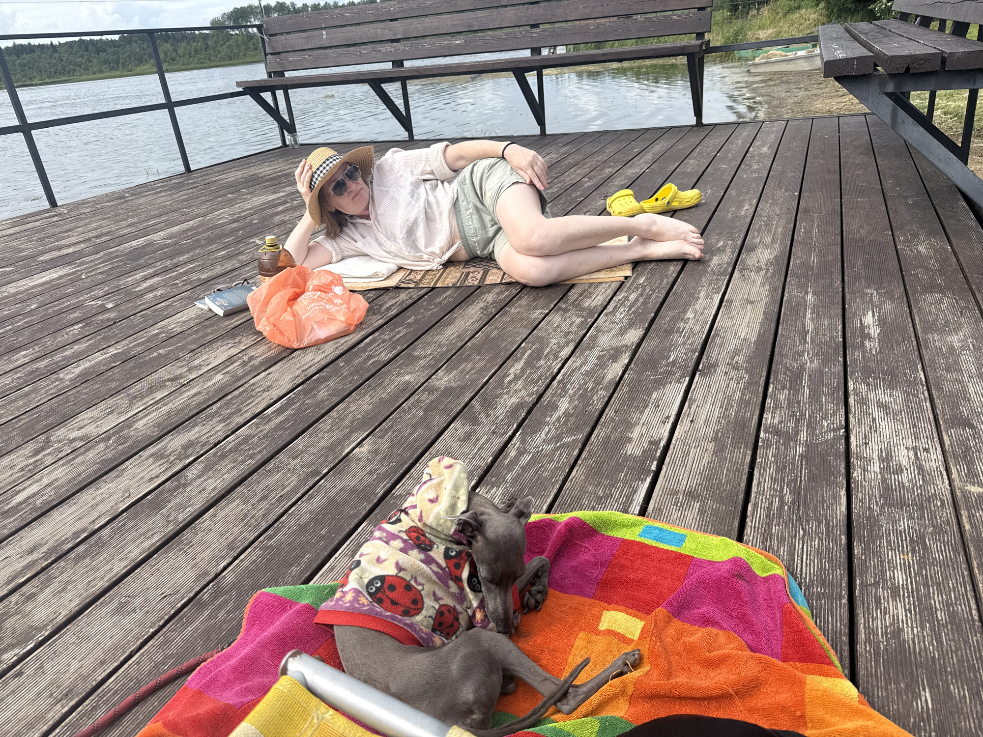 A person and a dog are on a wooden pier by a lake. The person, wearing a straw hat, sunglasses, a light-colored shirt, and shorts, is lying on their side on a mat. A bottle, an orange plastic bag, and books are near the person. A pair of yellow Crocs are on the pier. The dog, a greyhound wearing a hooded garment with ladybugs, is lying on a colorful striped towel. The pier has wooden planks and a railing. A wooden bench is visible in the background, as is a body of water with trees and a clear sky. The scene appears to be in Chikino, Russia.