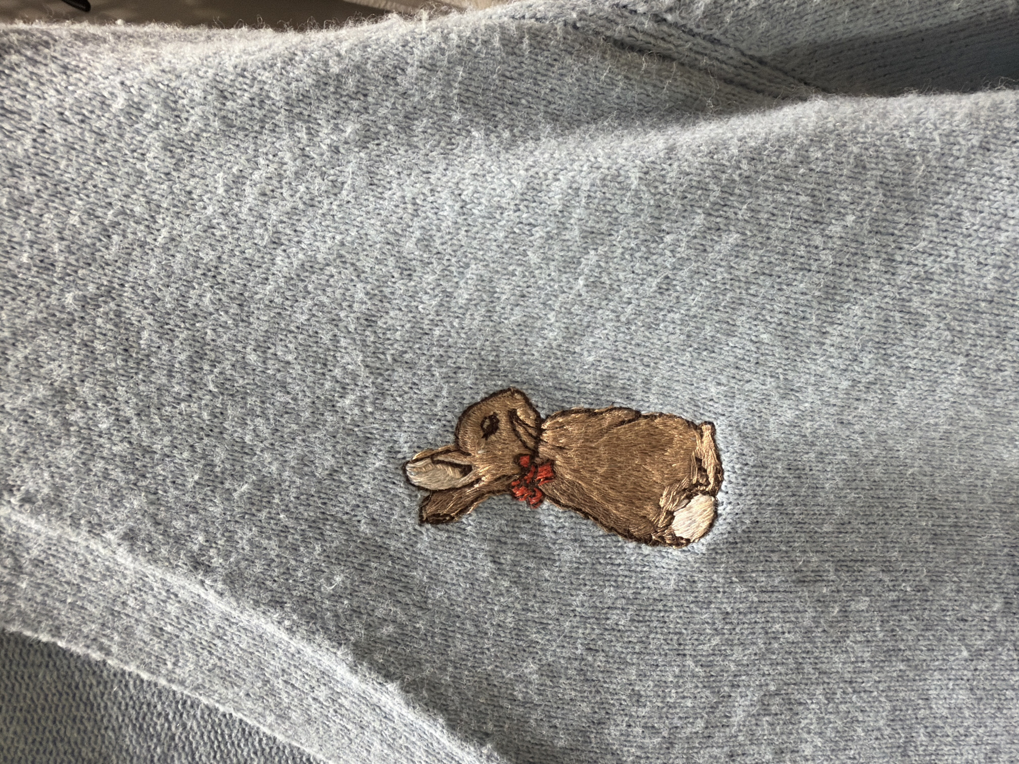 A close-up shot shows a light blue knitted sweater with a small embroidered brown bunny on the left side of the chest. The bunny is facing to the left, with its ears perked up and a small red bow tied around its neck. The sweater appears to be made of a soft, slightly fuzzy material, and the knit pattern is clearly visible. The background is out of focus but suggests the sweater is hanging or draped. The scene takes place indoors. There is no indication of the location in Denpasar, Indonesia.