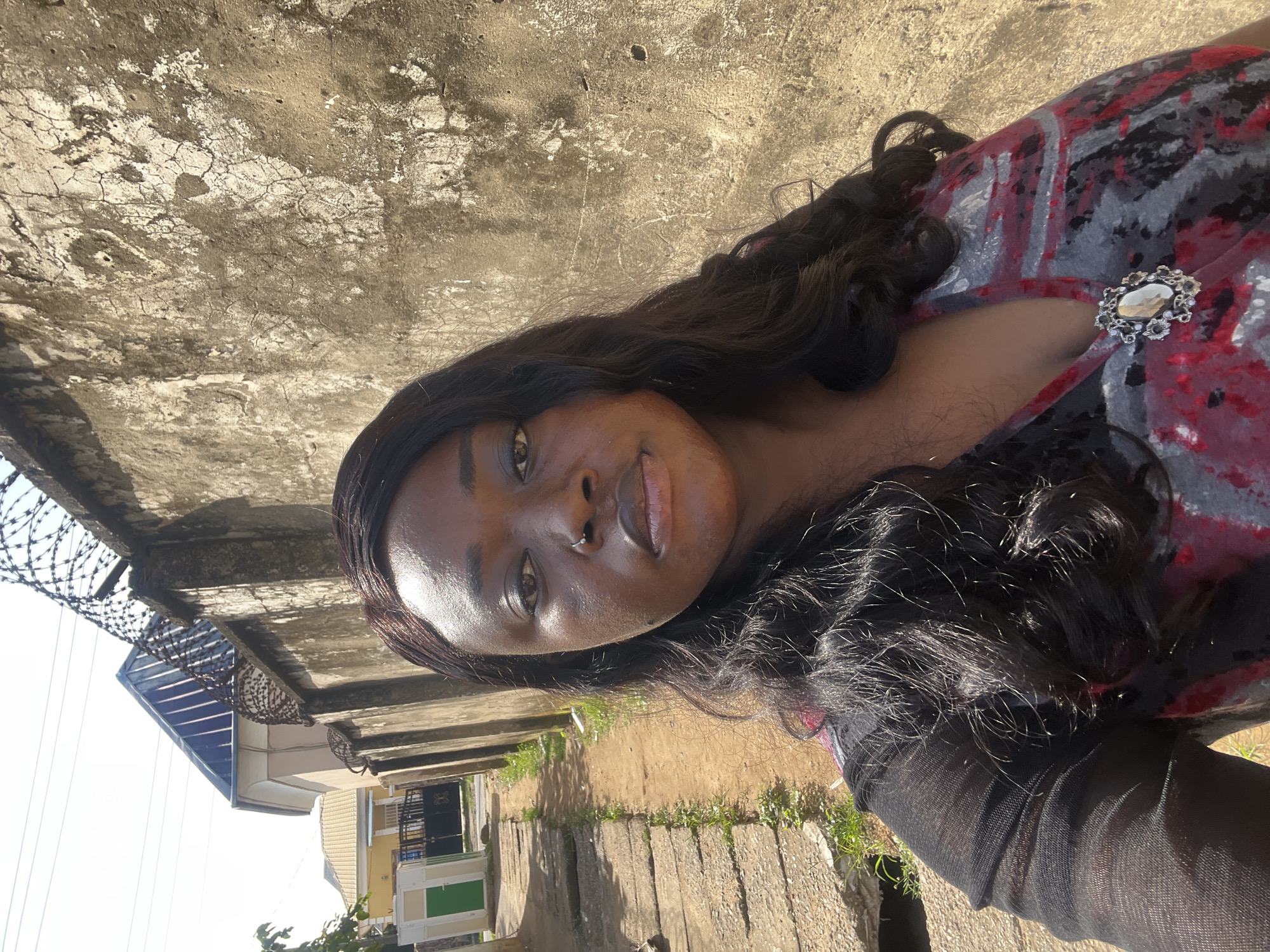 A woman is standing outside in what appears to be a residential area in Abuja, Nigeria. She is taking a selfie, facing the camera directly. She has dark, wavy hair and is wearing a dark-colored top with a red and gray abstract pattern. A decorative brooch is pinned to the neckline of her top. She has a nose ring. Behind her, a rough, textured wall runs along the side of a pathway. Barbed wire is visible along the top of the wall. In the background, houses with distinctive rooflines and a green and white structure can be seen. The ground is a dirt path with some concrete slabs. The overall lighting suggests it is daytime with bright sunlight.