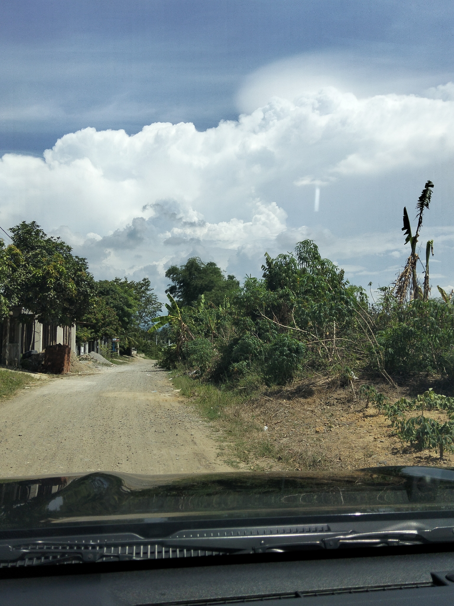 A dirt road winds through a rural area with lush vegetation on either side. On the left, a row of simple structures line the road, with trees providing shade. On the right, dense foliage and banana trees dominate the landscape. In the background, a vast expanse of sky is filled with dramatic, billowing white clouds against a blue backdrop. The image is taken from inside a vehicle, with the dashboard and windshield wipers visible in the foreground.