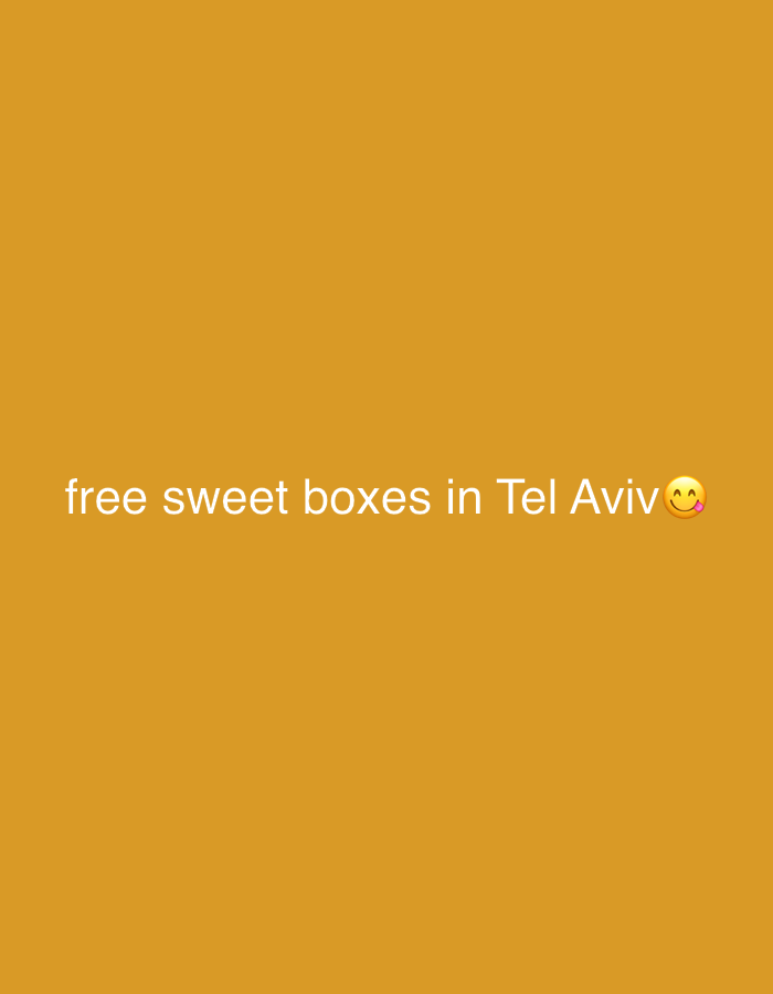 The image displays white text centered against a solid mustard yellow or deep orange background. The text reads: "free sweet boxes in Tel Aviv😋". An emoji depicting a face savoring food or licking its lips is placed at the end of the text. There are no other visible subjects, objects, or a discernible scene. The setting is a minimalist, single-color graphic with text overlay. The mention of "Tel Aviv" specifically indicates the location.