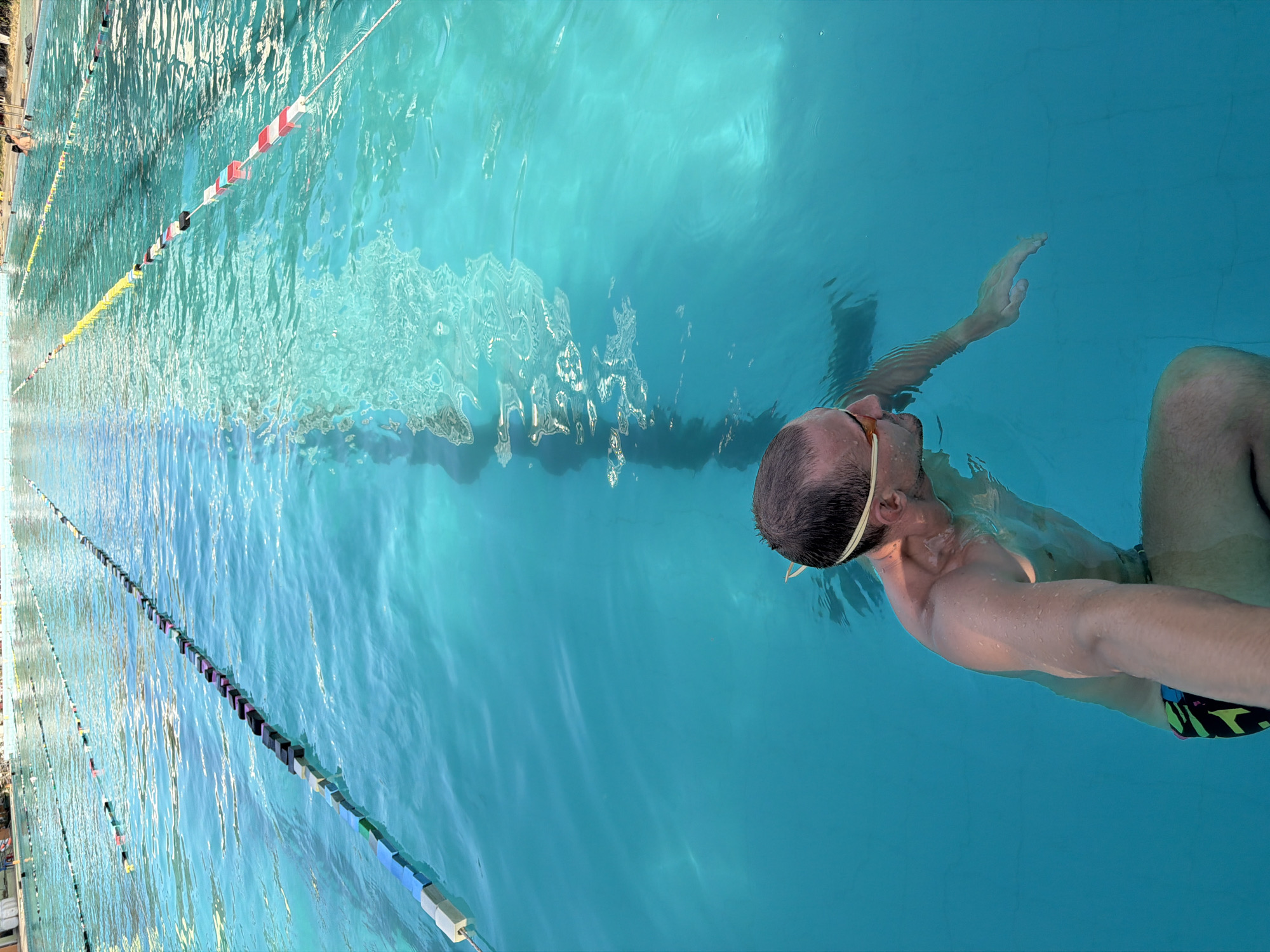 A high-angle shot shows a man swimming in an outdoor pool. The pool is demarcated into lanes by floating dividers. The man wears goggles and is partially submerged, with his right arm extended. The location may be in Nha Trang Ward, Vietnam, as suggested by the source.