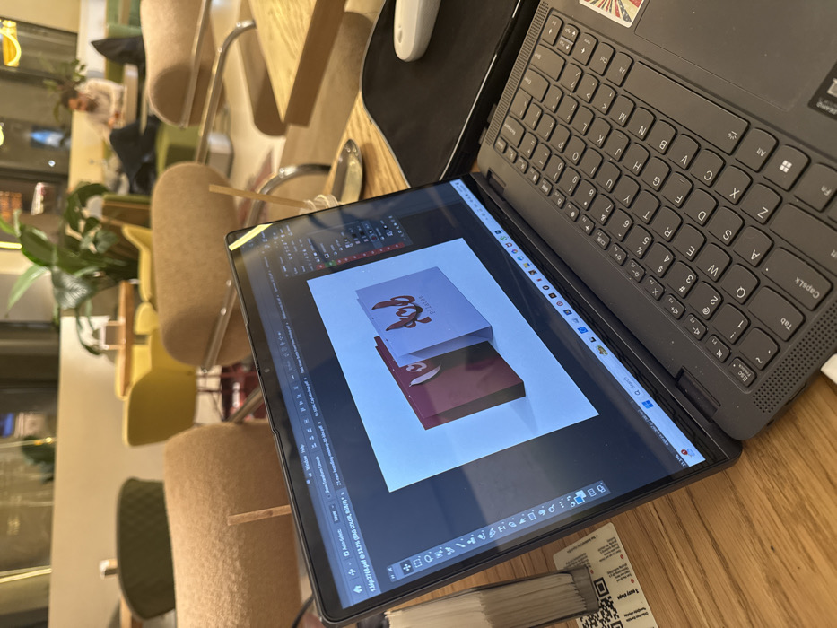 The image captures an indoor scene, likely a cafe or a co-working space, with a laptop and an external screen prominently displayed on a wooden table. The perspective is from above and to the right, looking down at the setup. The image is rotated clockwise, so the laptop screen appears on the left and the keyboard on the right.
The main focus is a dark gray or black laptop. The laptop is open, with its keyboard visible on the right side of the image. The keys are dark, and the layout appears to be QWERTY with additional function keys. Some keys have Arabic characters alongside English letters (e.g., Q, A, Z keys). A small, faded sticker, possibly an American flag design, is visible above the '6' key. A white USB cable is plugged into the right side of the laptop's base. To the right of the keyboard, there's a speaker grille.
Attached to the laptop, presumably as an external display, is a thinner tablet-like screen positioned to the left. This screen is displaying design software, likely Adobe Photoshop or a similar application, showing a mock-up of two rectangular package designs. The background of the design canvas is white. The top package is light gray with a stylized red design and the word "PLEASURES" underneath. The bottom package is a darker maroon or deep red with a similar stylized red design. The software interface shows toolbars on the left and bottom of the screen. The title bar at the top displays what appears to be a file name, "1.3.2766.pdf @ 31.3% (BAG COLOR, Granding...", indicating a PDF document being edited, possibly related to bag or packaging design.
To the upper left of the laptop (or upper right if oriented correctly), a light-colored computer mouse is visible on a dark mat.
The table beneath the laptop and screen is made of light-colored wood. In the foreground, below the laptop screen, a stack of white papers or small booklets is visible, with a QR code partly discernible on the top sheet.
The background of the scene shows a soft-focused cafe environment. There are several chairs with beige upholstered seats and backs, some with wooden frames, and others with yellow or dark green upholstery. Green plants are scattered around, adding to the cafe aesthetic. Further in the background, a person is faintly visible, seated at a table, appearing blurry. The overall lighting suggests an interior space with possibly soft artificial lighting or natural light filtering in.
Visible text from the OCR:
* On the screen: "1.3.2766.pdf @ 31.3% (BAG COLOR, Granding...", "PLEASURES"
* On the keyboard: "Esc", "Tab", "CapsLk", "Shift", "Fn", "Alt", Windows logo key, "Ctrl", "Sch", "del", "Pilates" (on a keycap), "Q", "W", "E", "R", "T", "Y", "U", "I", "O", "P", "A", "S", "D", "F", "G", "H", "J", "K", "L", "Z", "X", "C", "V", "B", "N", "M", "<", ">", "/", "?", "Alt Gr", "+", "Enter", "Backspace", "Spacebar", number row keys "1" through "0" with symbols "#", "$", "%", "^", "&", "*", "(", ")". Some Arabic characters are present on keys like 'Q', 'A', 'Z', 'W', 'S', 'X', 'E', 'D', 'C', 'R', 'F', 'V', 'T', 'G', 'B', 'Y', 'H', 'N', 'U', 'J', 'M', 'I', 'K', 'O', 'L', 'P'.