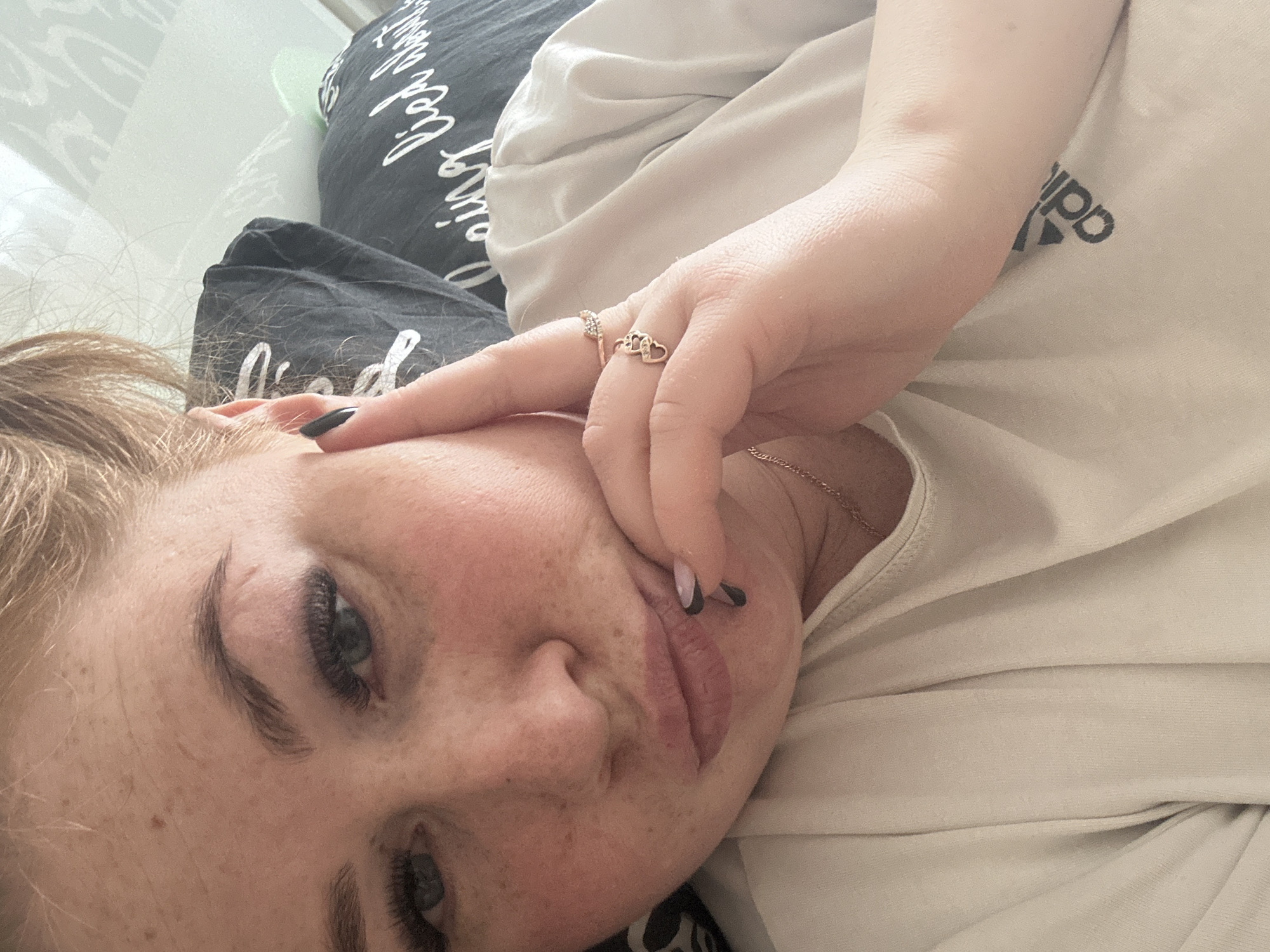 A woman with fair skin, freckles, and light brown hair is lying down. She has black nail polish on her fingernails. The woman wears rings on her fingers. She is wearing a light-colored t-shirt with the Adidas logo on the front and is lying on a gray pillow. The image was not taken in Chaikovsky, Russia, so I cannot provide any information about the location of the image.