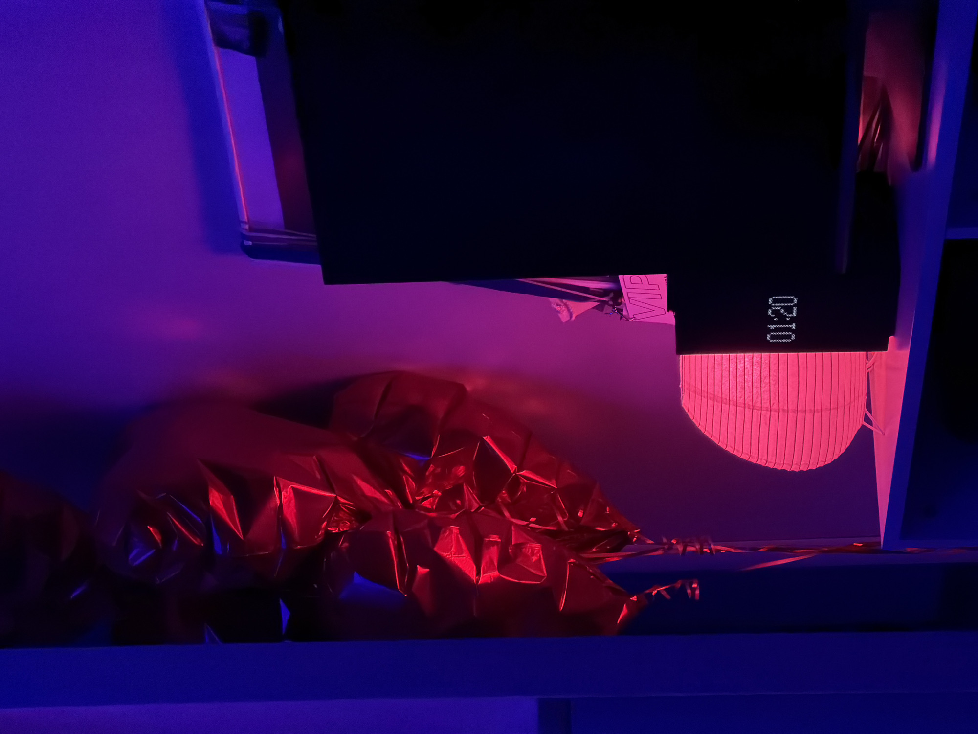 The image shows a scene indoors, illuminated by predominantly red and blue lighting. A crumpled, reflective material, possibly foil or metallic fabric, is present in the foreground. Above this, a dark rectangular object is visible, with a digital display showing "01:20." A cylindrical, vertically ribbed object, possibly a lantern or lampshade, appears lit in red. The backdrop is a wall with a gradient of purple and blue. A "VIP" tag is partially visible. The location is unknown, but the prompt asks to assume it's in Krasnoyarsk, Russia.