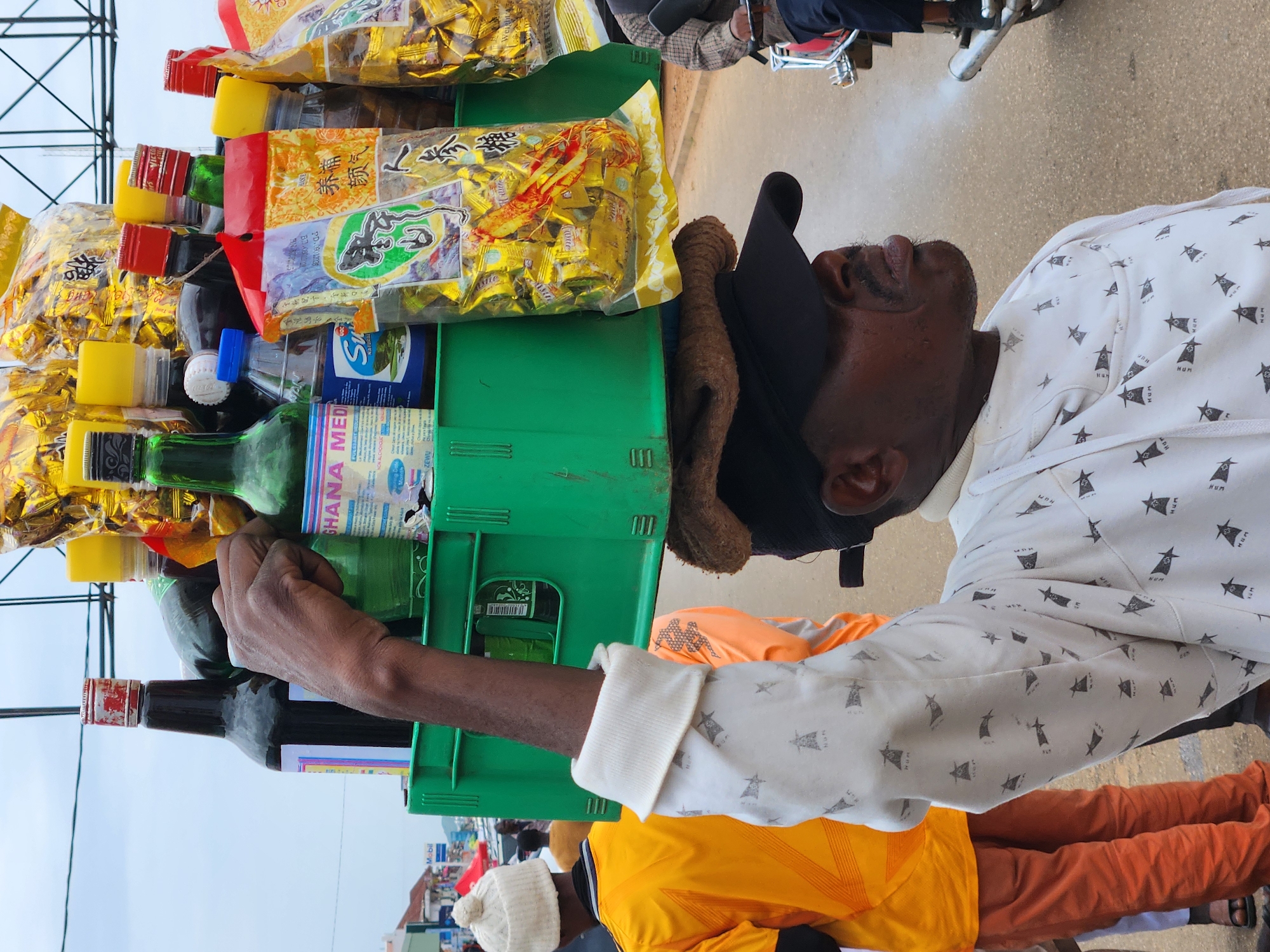 A man balances a green plastic crate filled with bottles and packets of goods on his head in Jos, Nigeria. The crate contains various items, including a large dark bottle with a red and white cap, a green bottle, a bottle with a blue cap and a label reading "Sun Natural", and several golden packets with Chinese writing. The man is wearing a white long-sleeved shirt with a repeating pattern of stylized figures and a black cap. He is looking to the right, and his mouth is open as if he is exhaling. In the background, other people are visible, including a person in a bright yellow shirt and orange pants, and another person in an orange shirt. A motorcycle is also visible to the right. The scene appears to be taking place on a street with a paved surface.