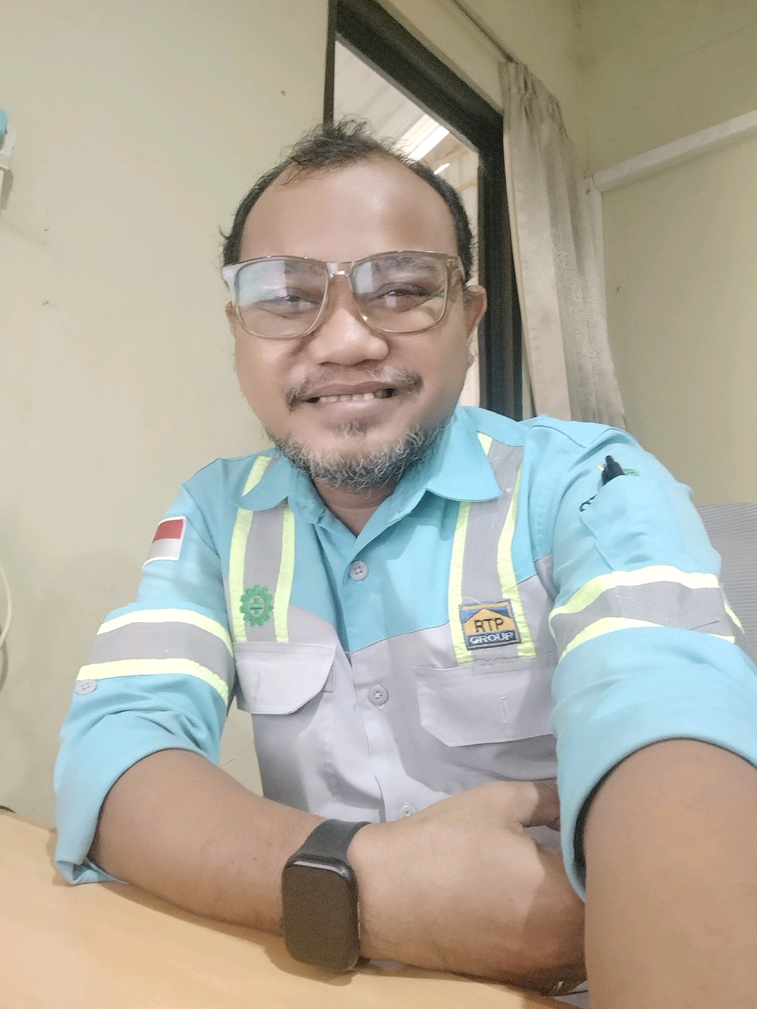 The image is a selfie of a man in what appears to be a workplace environment. The man is wearing a light blue collared shirt with grey and yellow reflective stripes across the chest and sleeves. He has a badge on his left chest with the Indonesian flag and another on his right chest with the RTP GROUP logo. He is also wearing glasses and a black smartwatch. The background shows a plain wall and a window with beige curtains. The man is smiling at the camera, with his hands resting on the table in front of him. The scene is likely indoors, possibly an office or a workshop in Maburai, Indonesia.