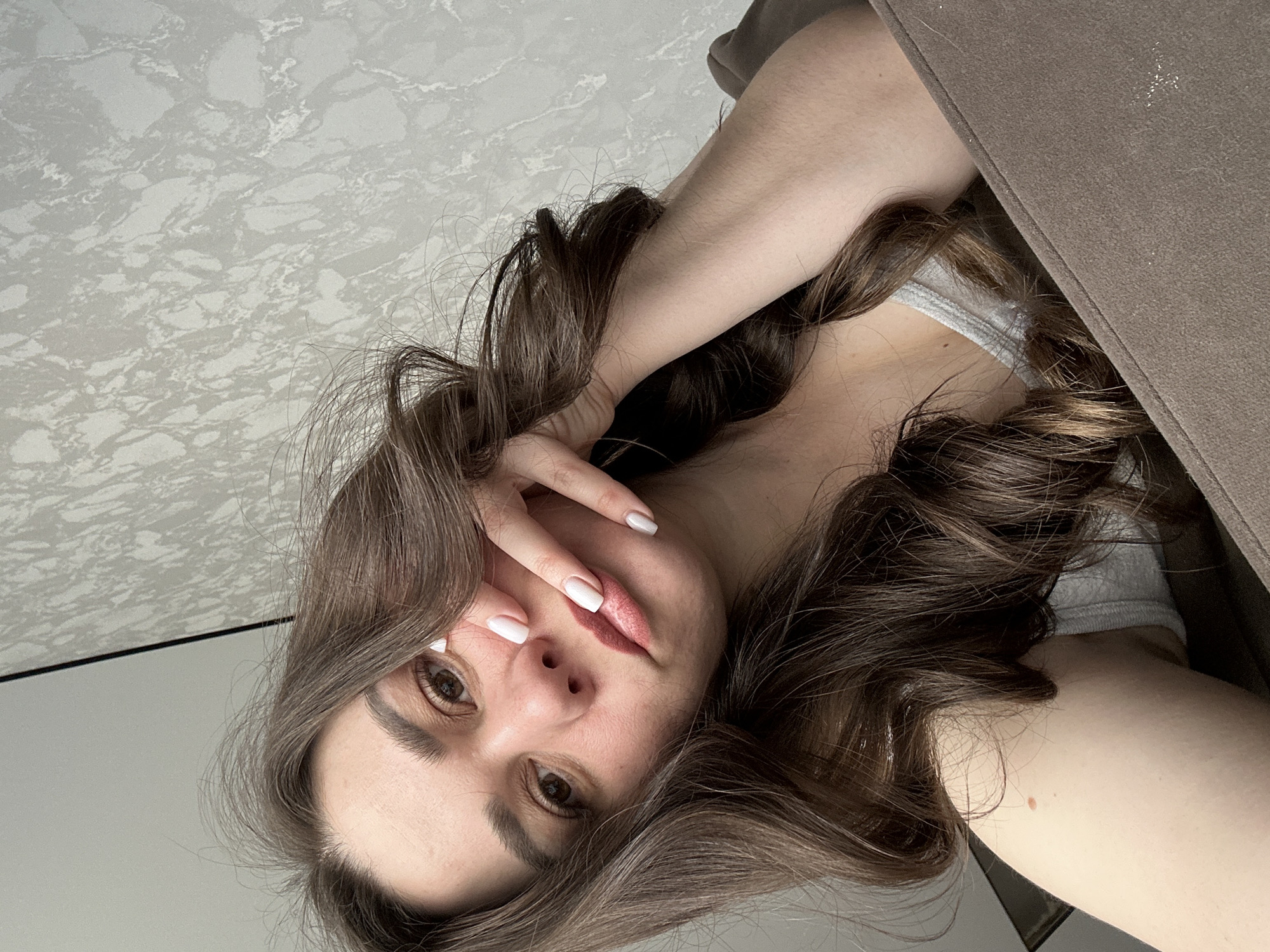 A woman is positioned in a low-angle selfie indoors. She has long, dark, curly hair, fair skin, and white painted fingernails. Her left hand is raised and partially covers her face. The background includes what appears to be a patterned white surface, possibly a countertop or wall panel, and a brown upholstered object, like a piece of furniture. No location data is discernable from the image to associate it with Chaikovsky, Russia, or any specific city.